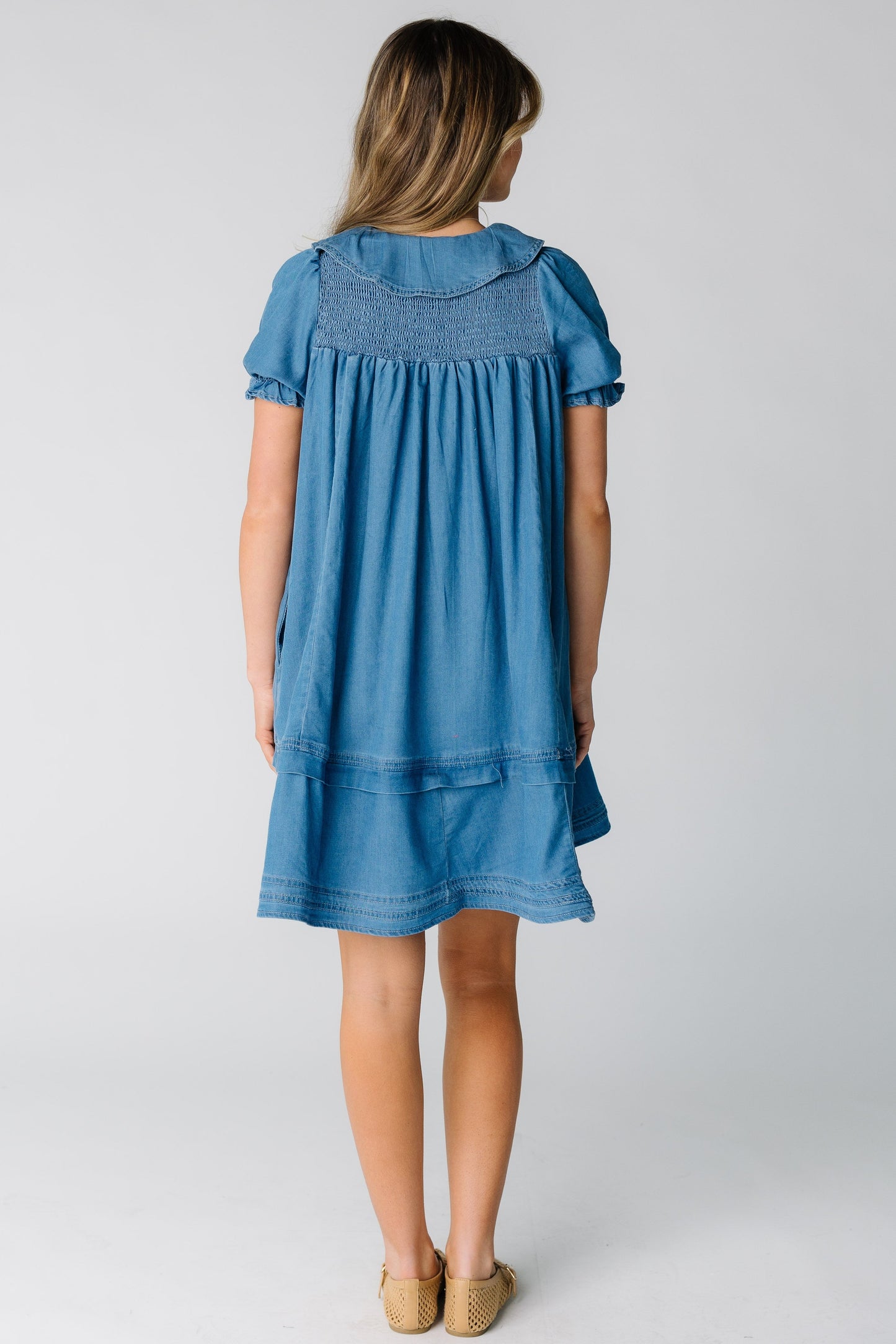 Back view of a woman wearing a knee length denim dress with a smocked back yoke and short puff sleeves.