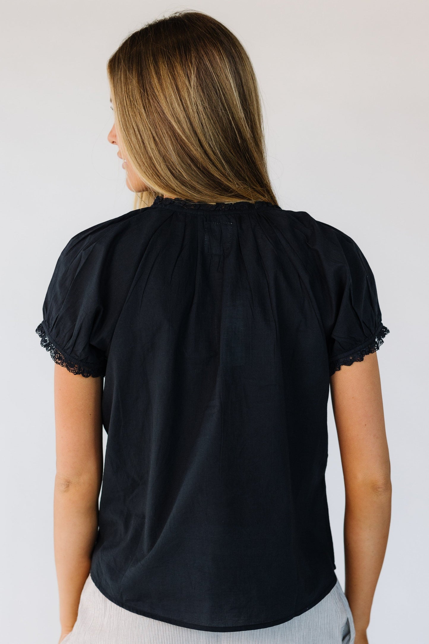Back view of a woman wearing a black woven blouse with a lace trimmed round neckline ad short puff sleeves with a lace hem.
