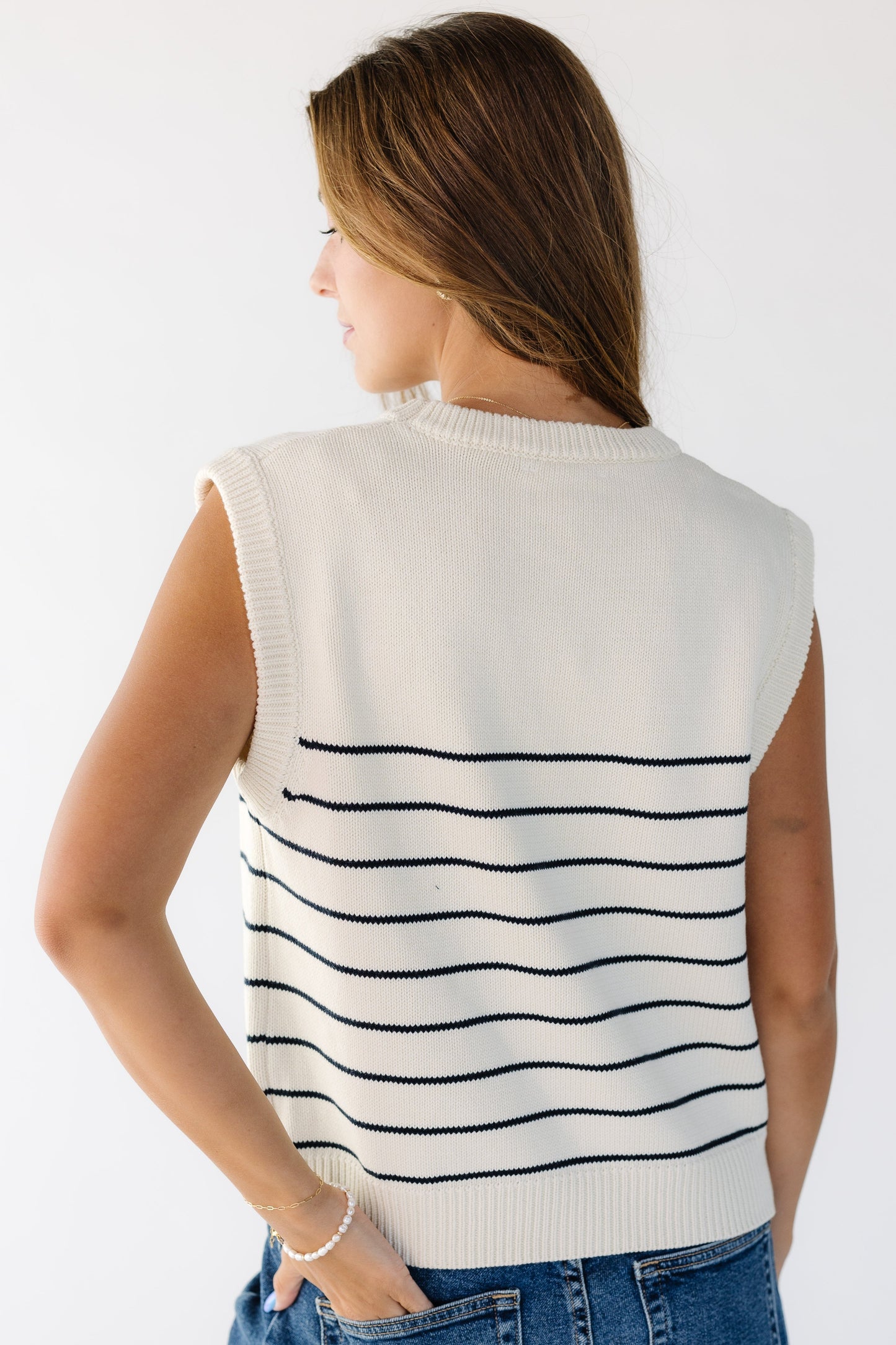 Back view of a young woman wearing a natural pullover sweater vest with a crew neckline and thin black stripes.
