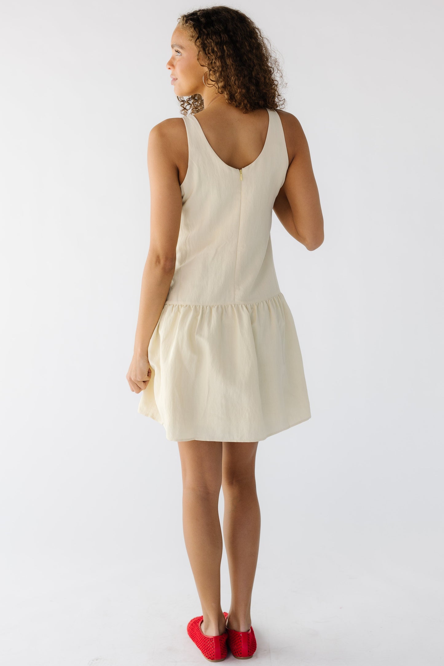 Back view of a teenage girl wearing a sleeveless light yellow dress with a gathered skirt and a back scoop neckline.