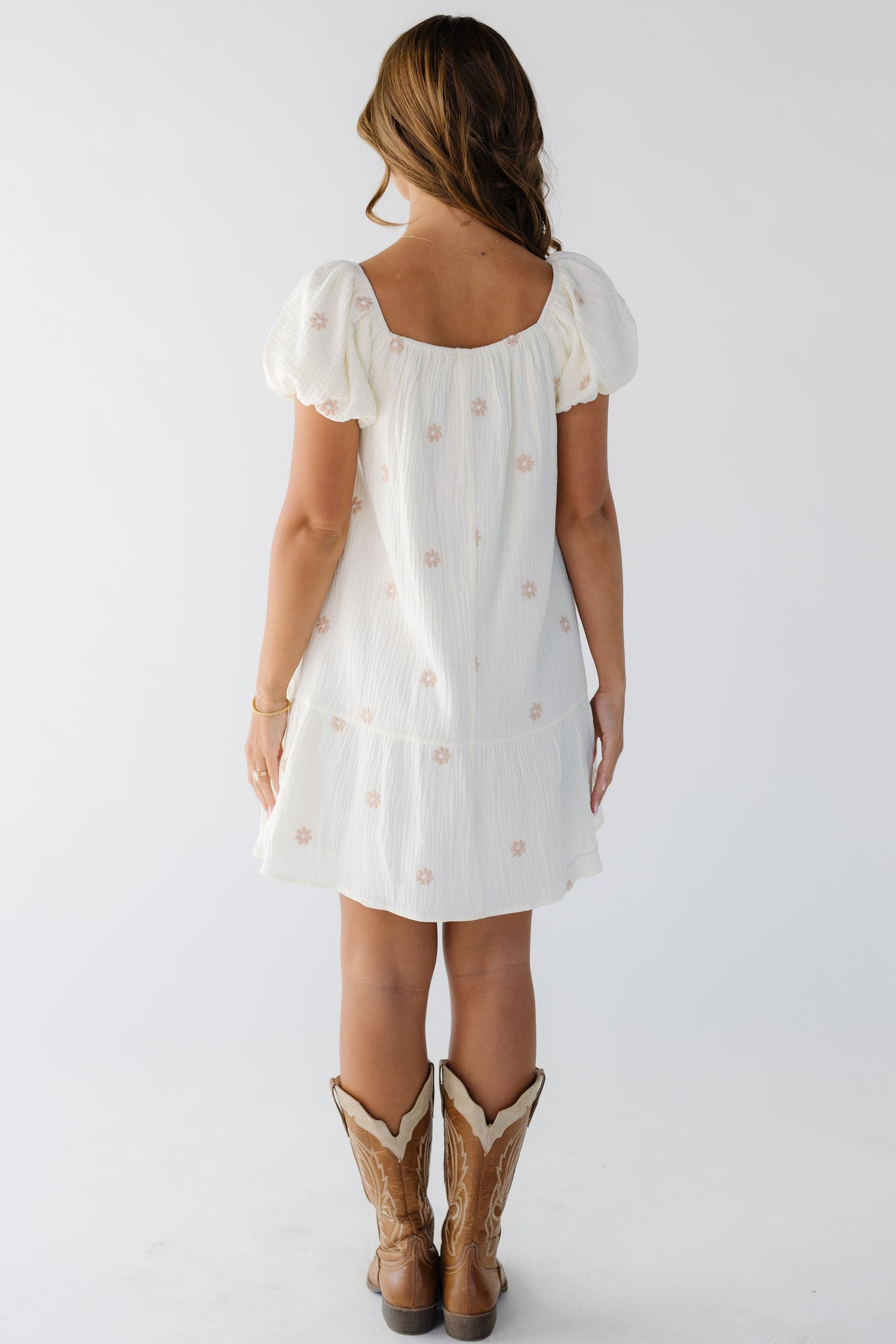 Back view of a woman wearing a short loose fitting ivory dress with pink floral embroidery with a square neckline and short puff sleeves.