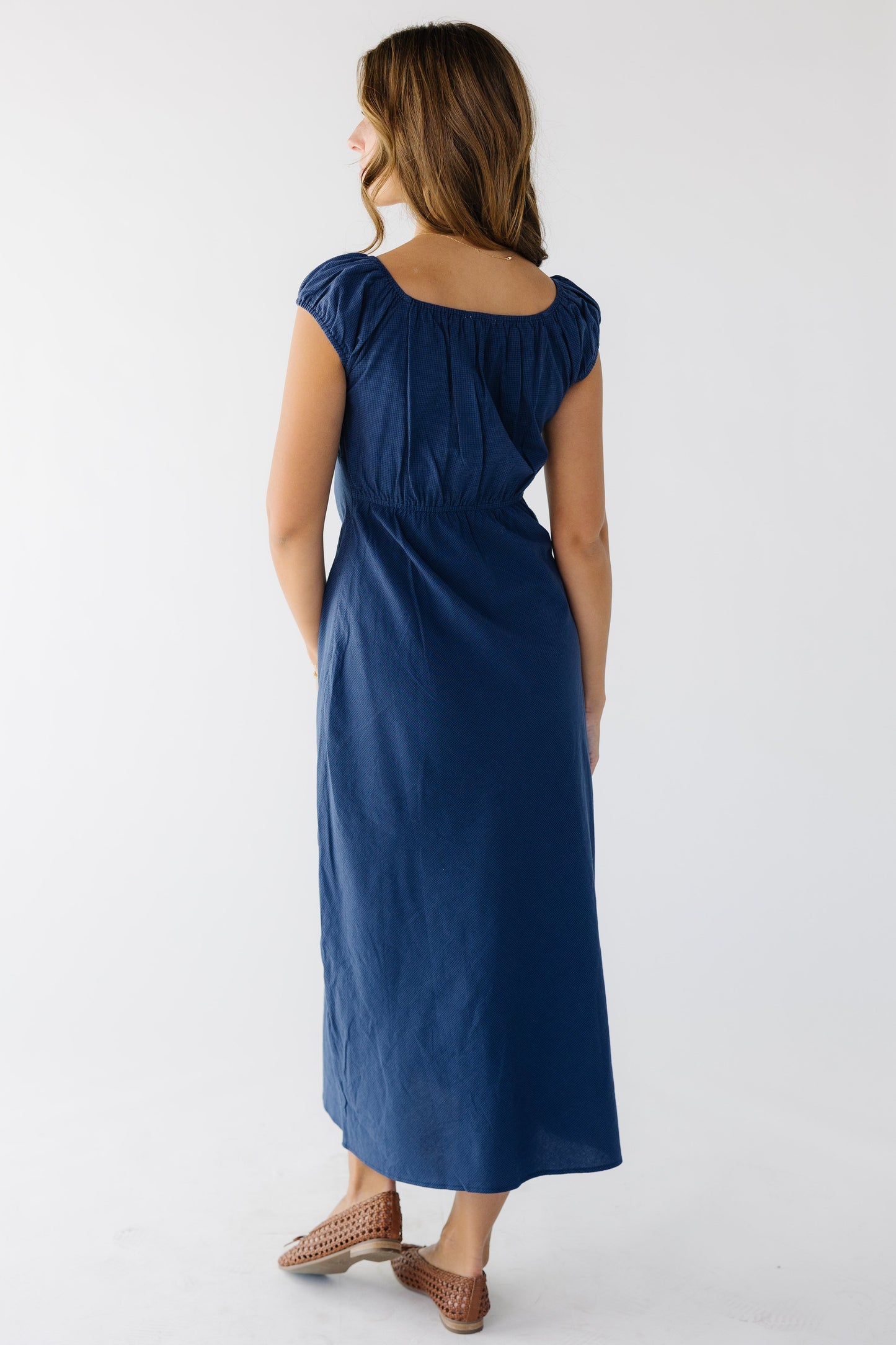 Back view of a woman wearing a navy A-line midi dress with an elastic back waist with a scoop neckline and short puff sleeves.