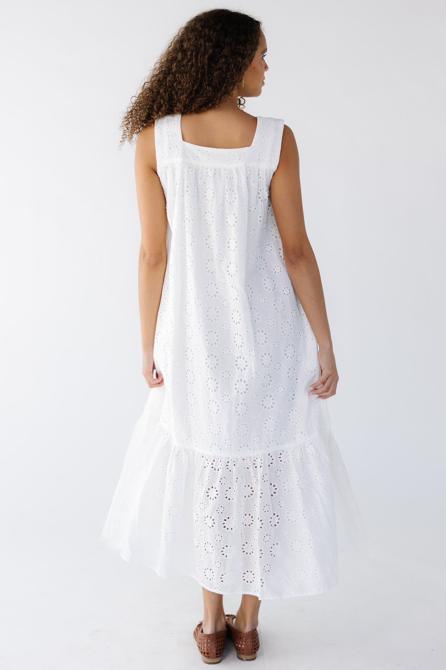 Back view of a young woman wearing a white sleeveless midi-maxi dress with a wide bottom ruffle, and a square neckline.