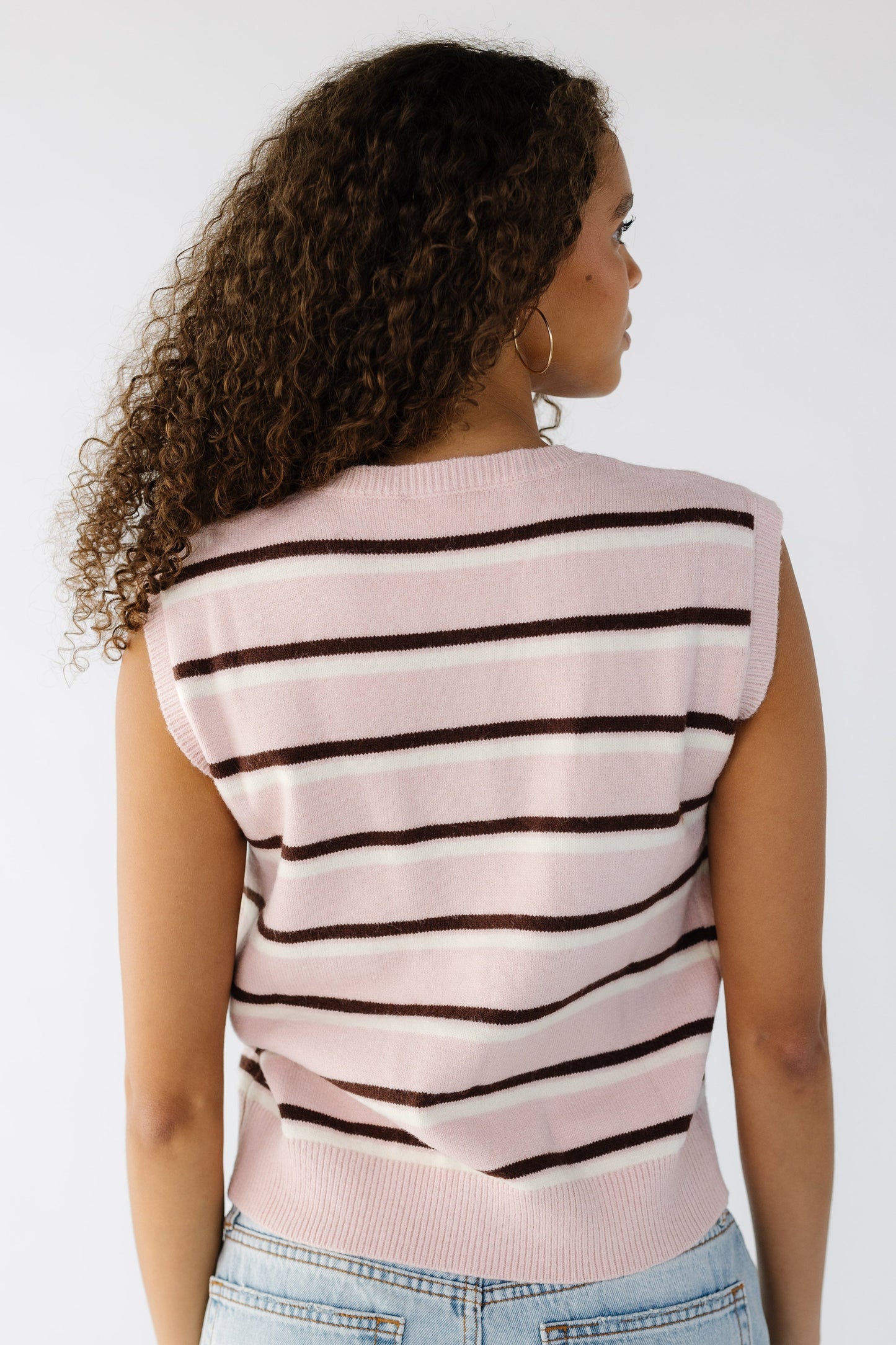 Back view of a teen girl wearing a pink, brown and cream stripe sleeveless sweater vest.