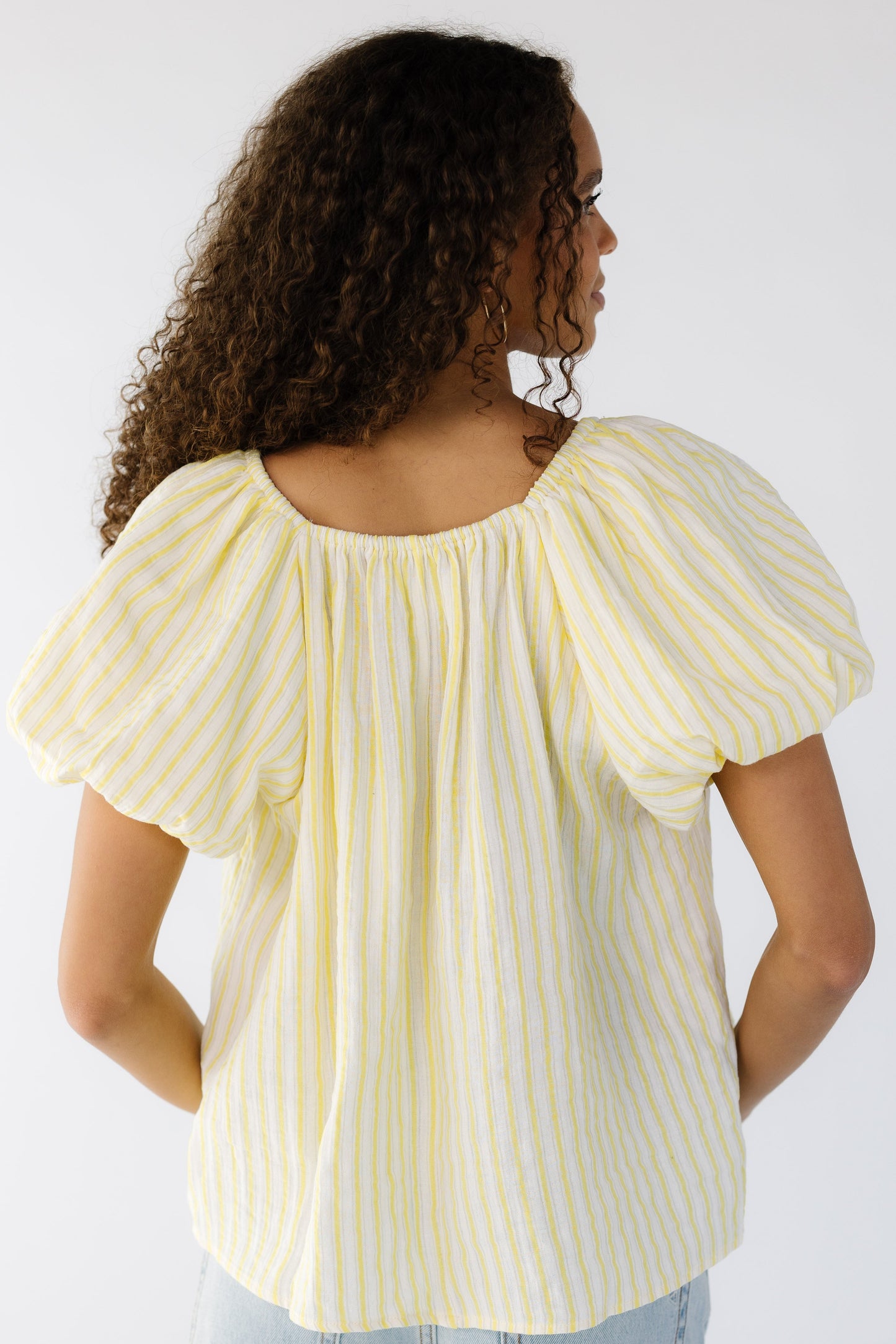 Back view of a teen girl wearing a yellow and white vertical stripe blouse with an elastic-square neckline with large balloon sleeves.