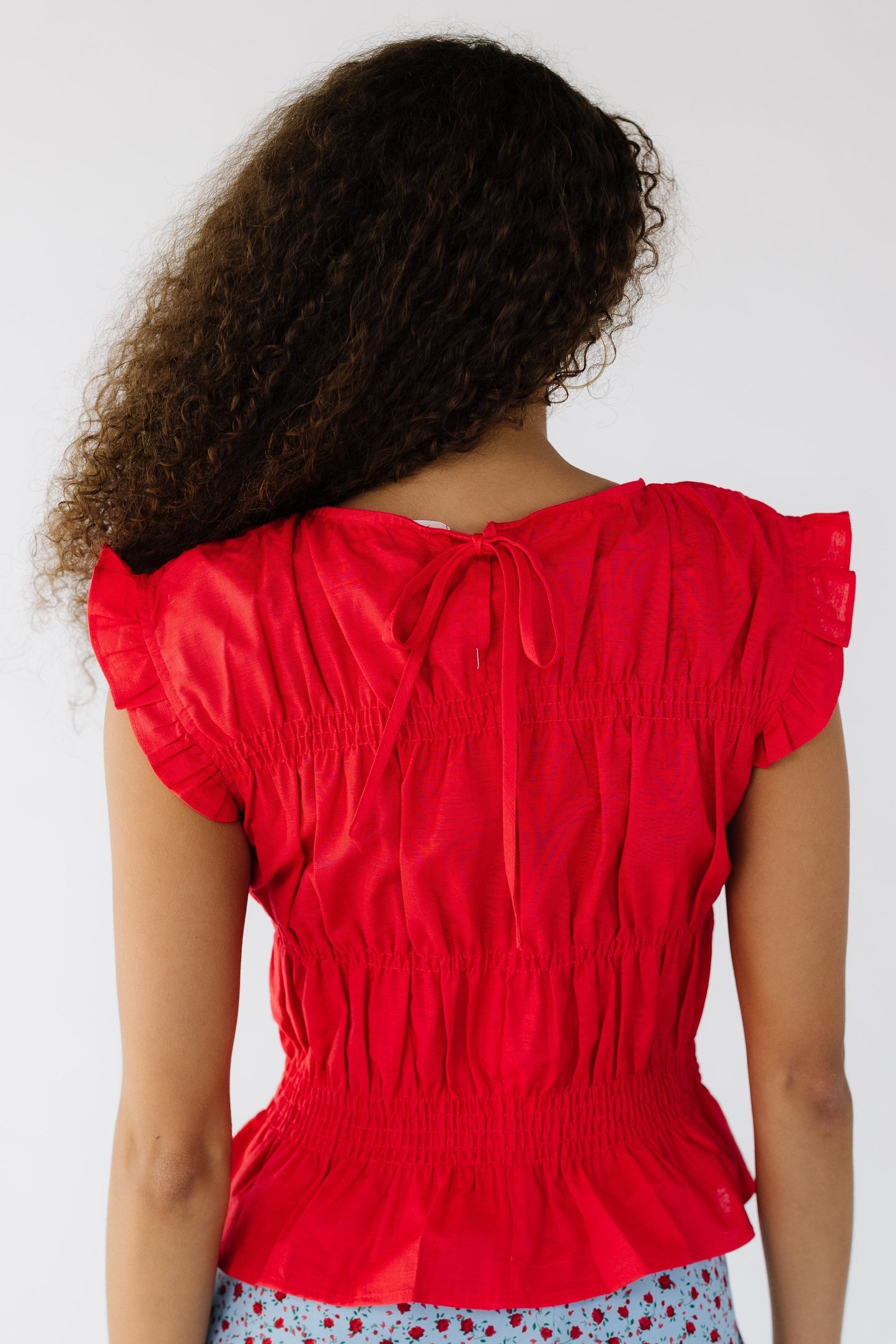 Back view of a young woman wearing a red sleeveless blouse with clustered rows of smocking.