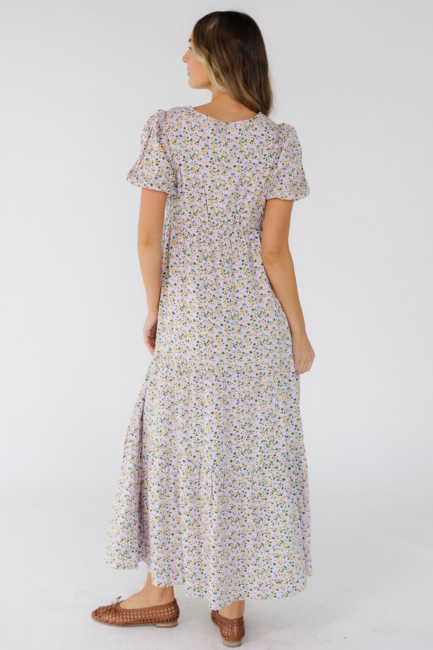 Back view of a woman wearing a lavender midi-maxi dress with puff sleeves, empire elastic waist and a loose tiered skirt.