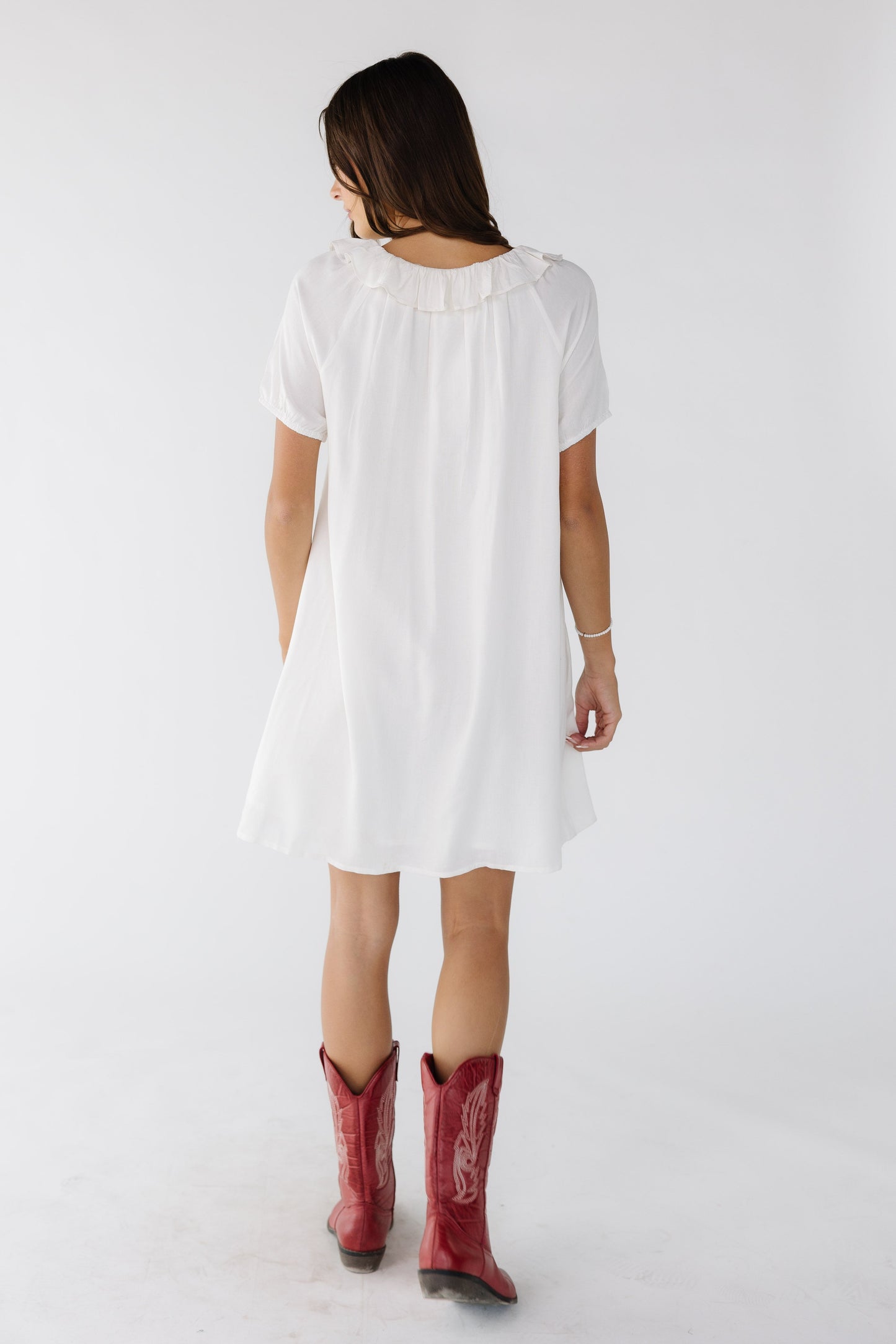 Back view of a woman wearing a white above the knee dress with a round ruffled neckline and short sleeves.