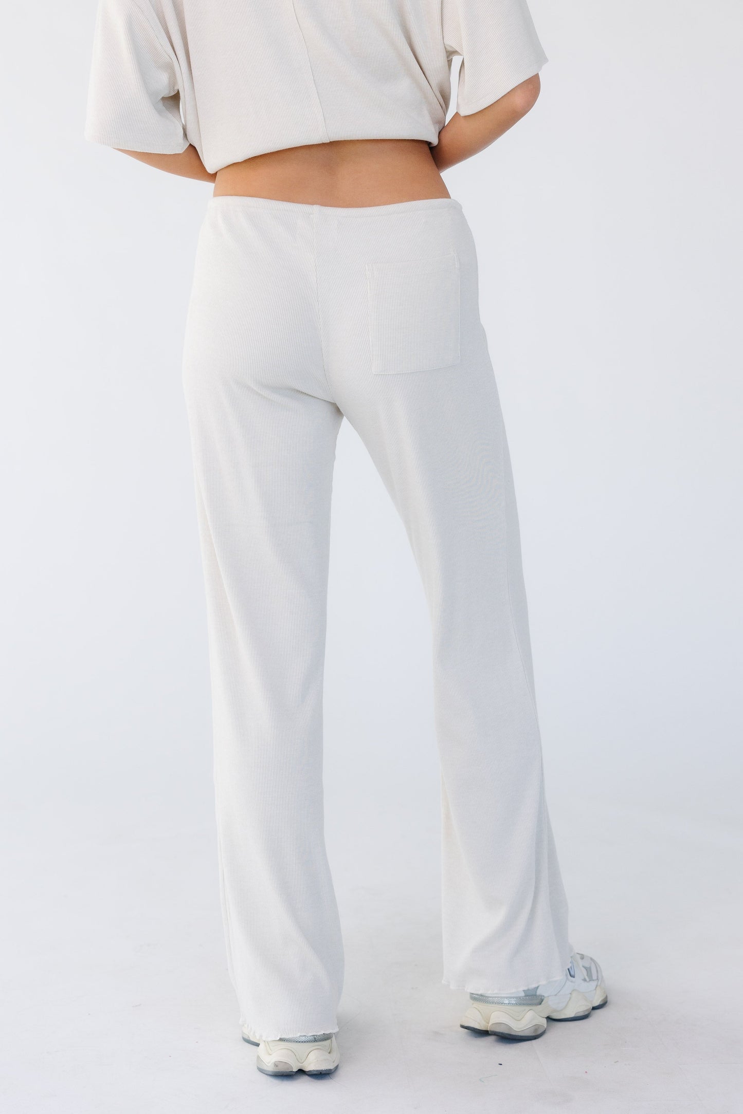Back view of a young woman wearing a pair of lounge pants that are close fitting through the waist & hip with a wide cut leg in light beige.