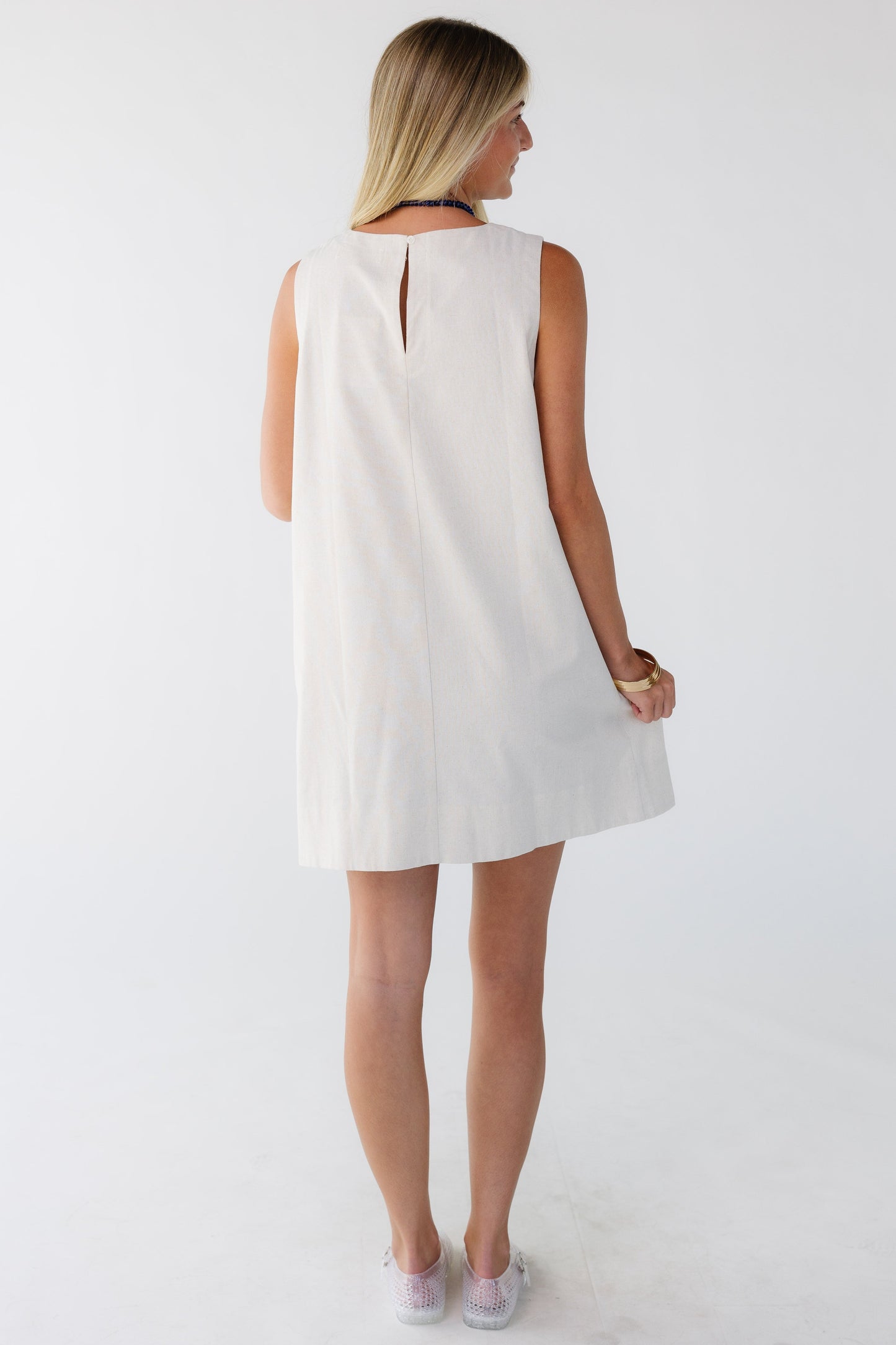 Back view of a woman wearing a cream loose fitting sleeveless shift dress.