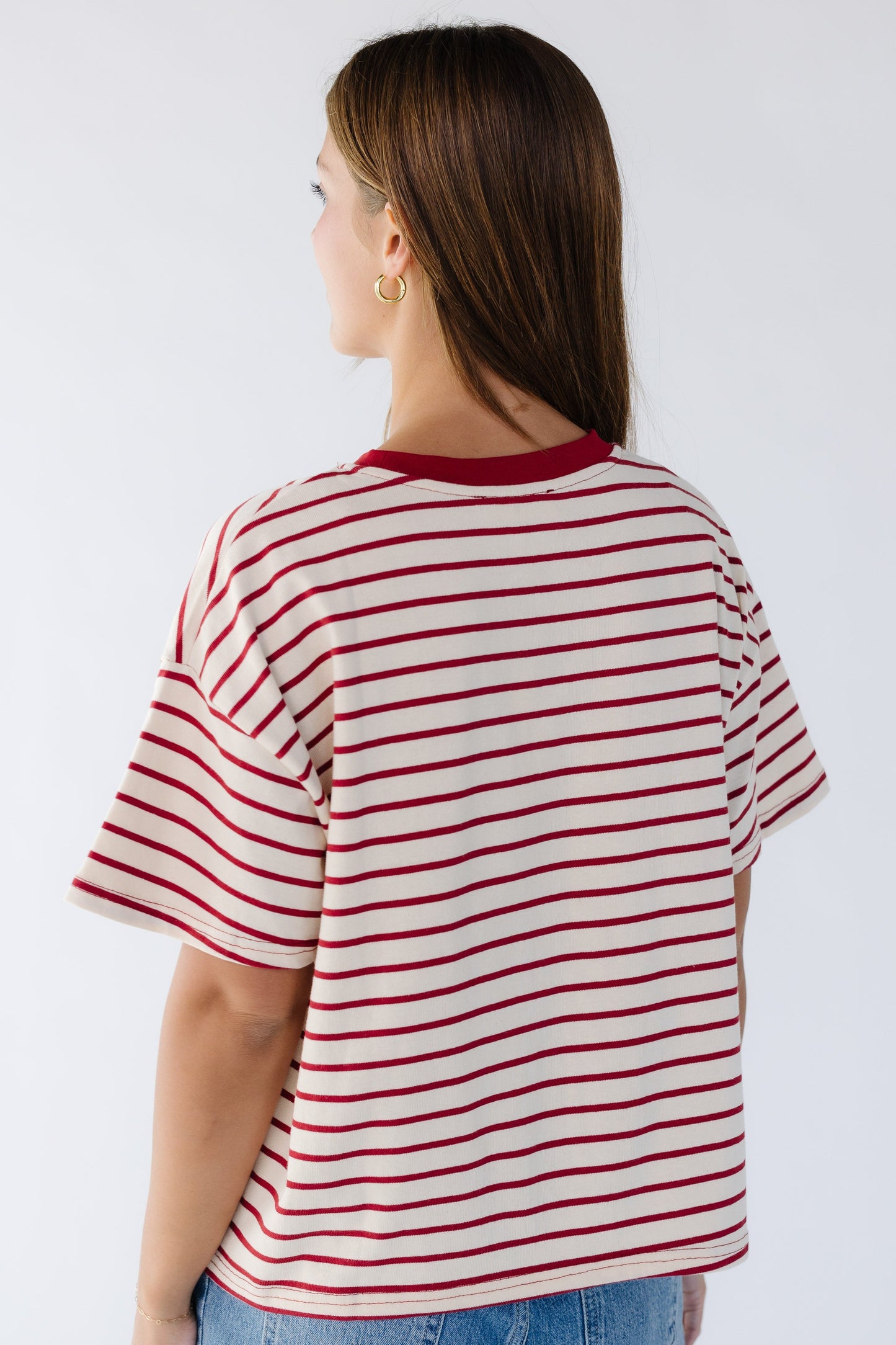 Back view of a woman wearing a cream and red stripe loose fitting tee with short sleeves and a red ribbed crew neckline.
