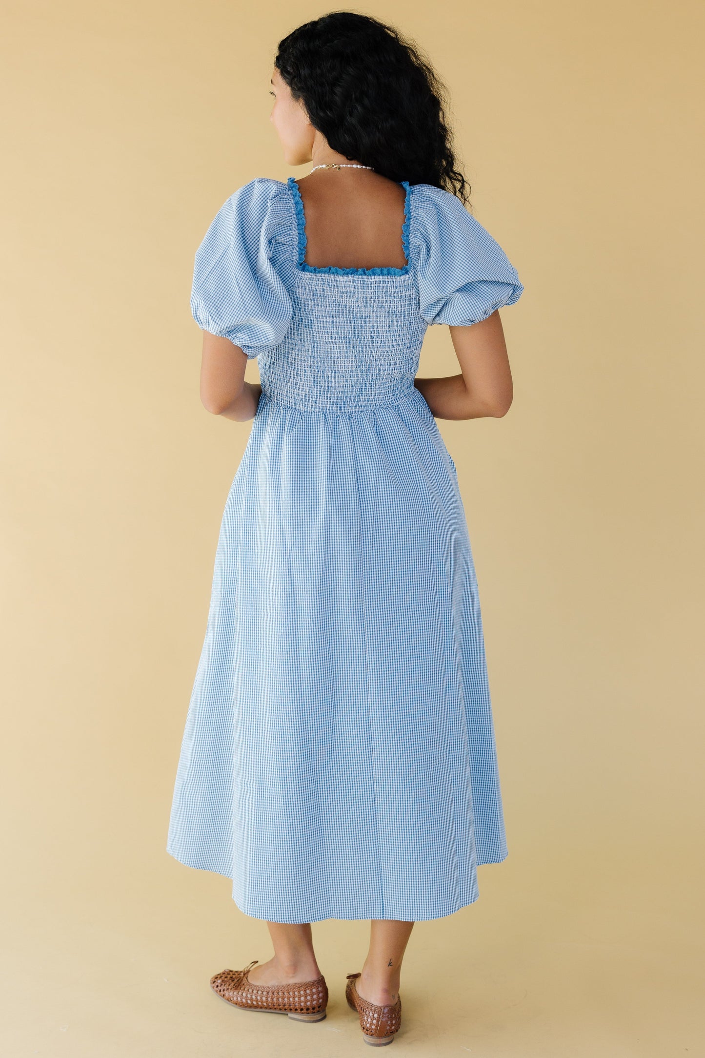 Back view of a young woman wearing a blue & white gingham midi dress with balloon sleeves and a lace trimmed square neckline.