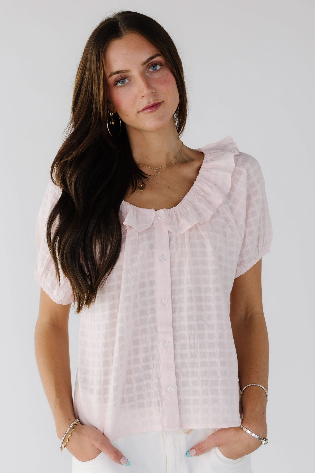 Young woman wearing a light pink, plaid textured button down top with short sleeves and a ruffled collar.