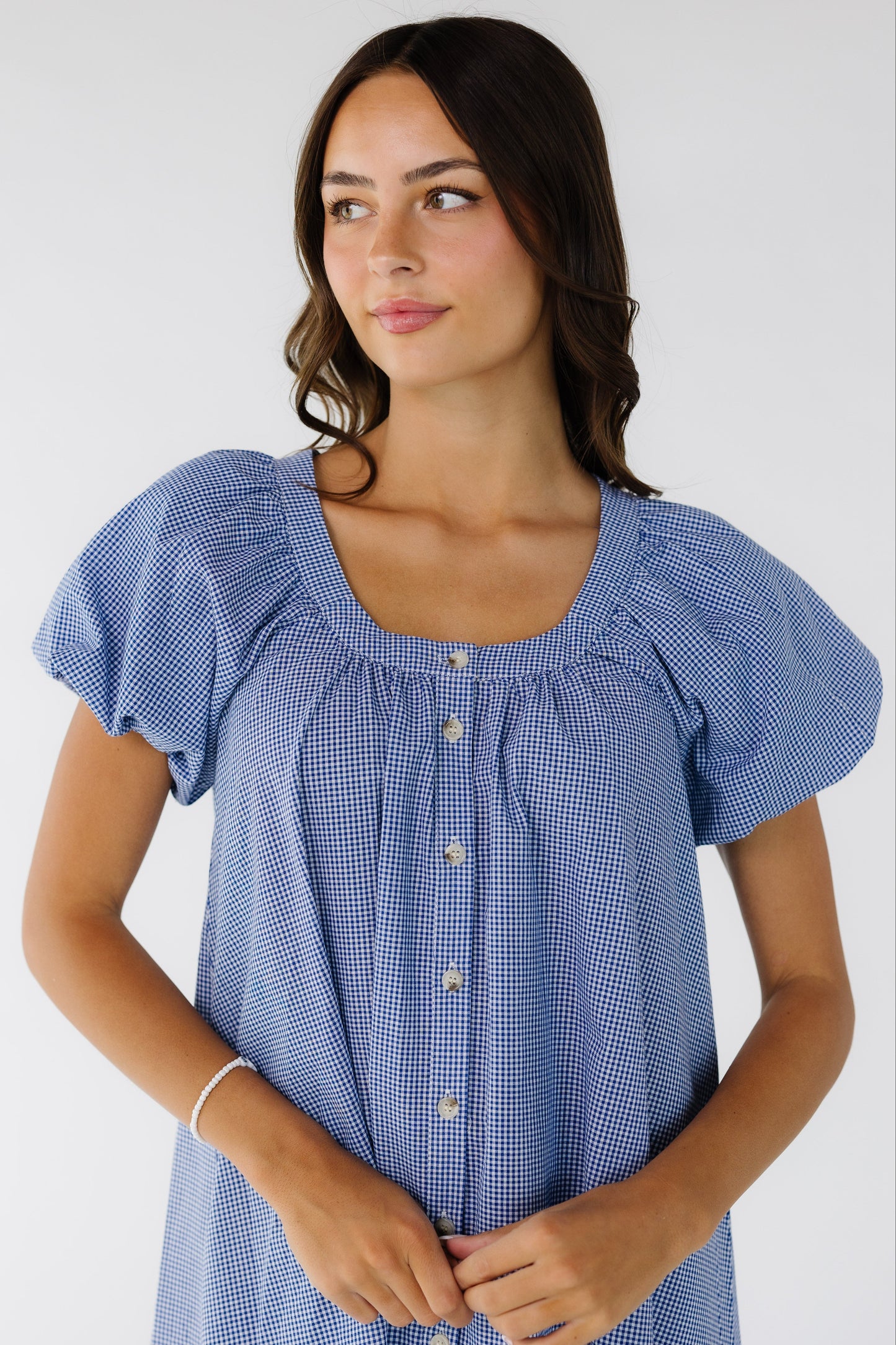 Close up view of a young woman wearing a navy & white gingham button down dress with balloon sleeves and a scoop neckline.