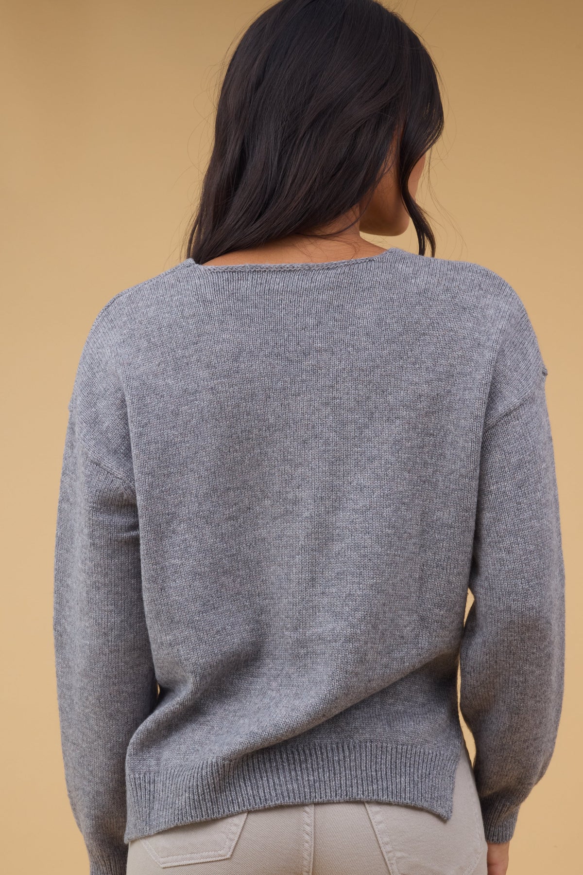 Back view of a woman wearing a grey pullover sweater with long sleeves and side slits.