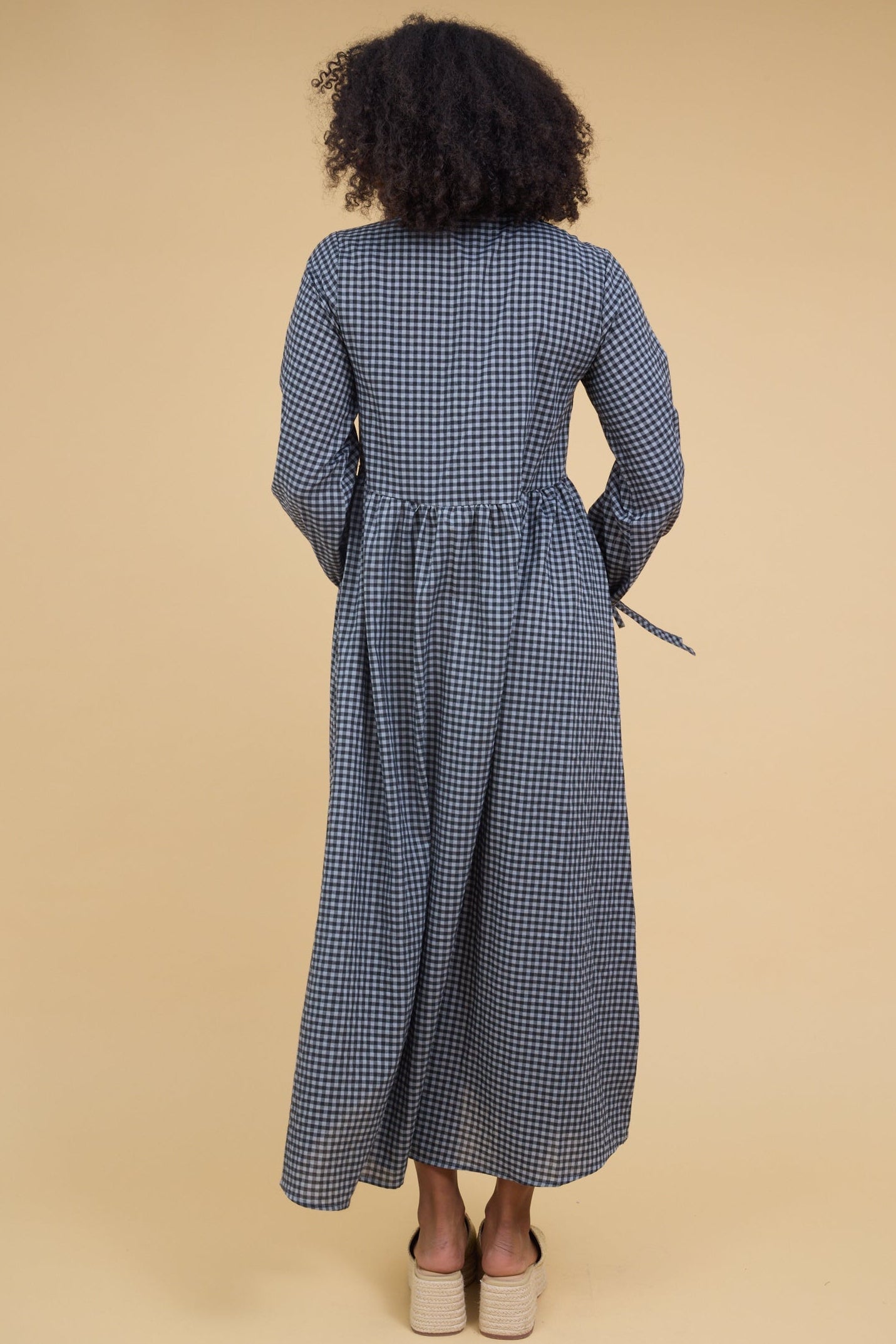 Back view of a woman wearing a blue gingham midi dress with long sleeves and a long full, gathered skirt.