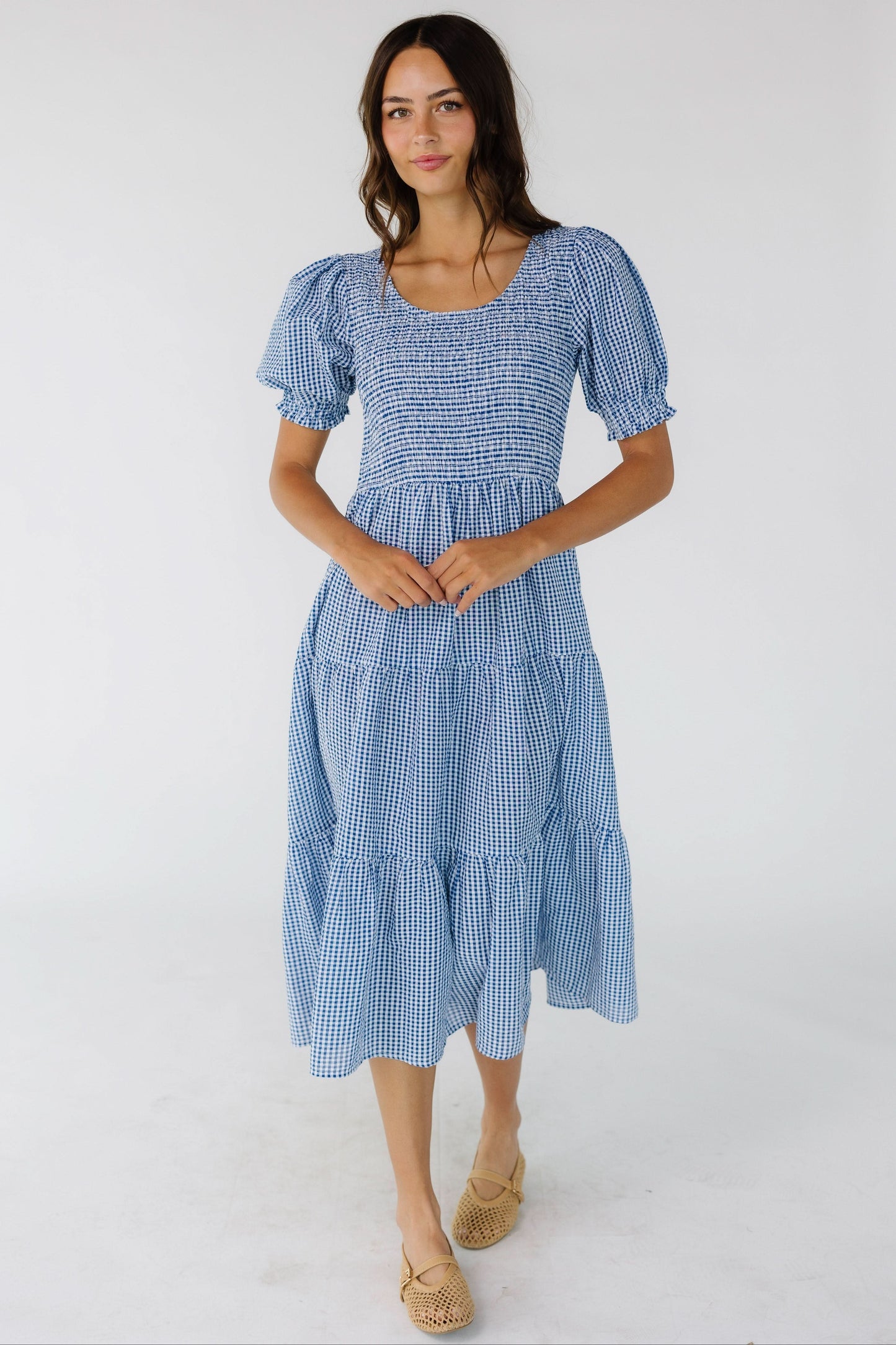 Young woman wearing a blue gingham midi dress with short puff sleeves, a scoop neckline and multiple gathered tieres.
