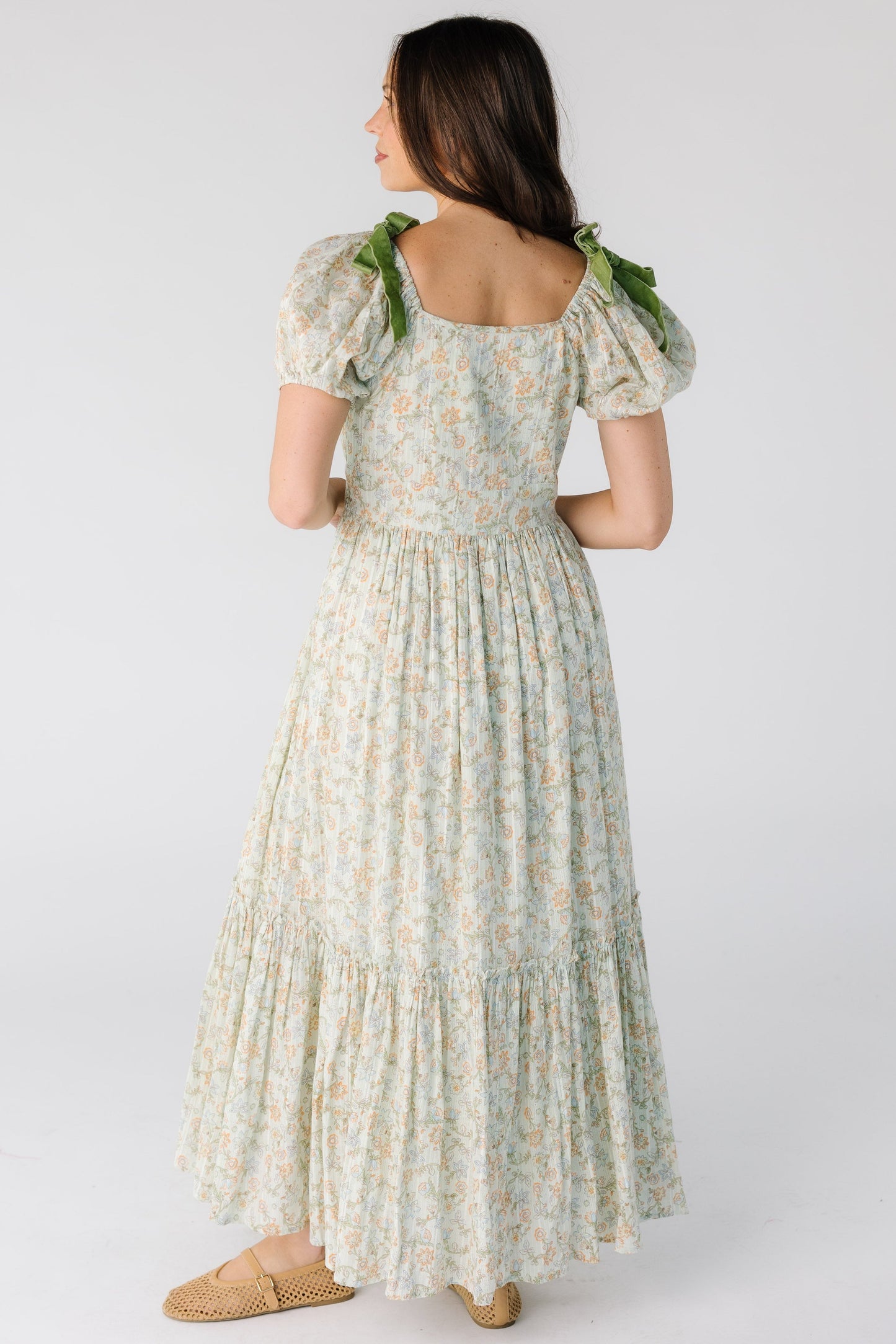 Back view of a woman wearing a green floral midi-maxi dress with a close fitting bodice and waist with full gathered skirt and short puff sleeves.