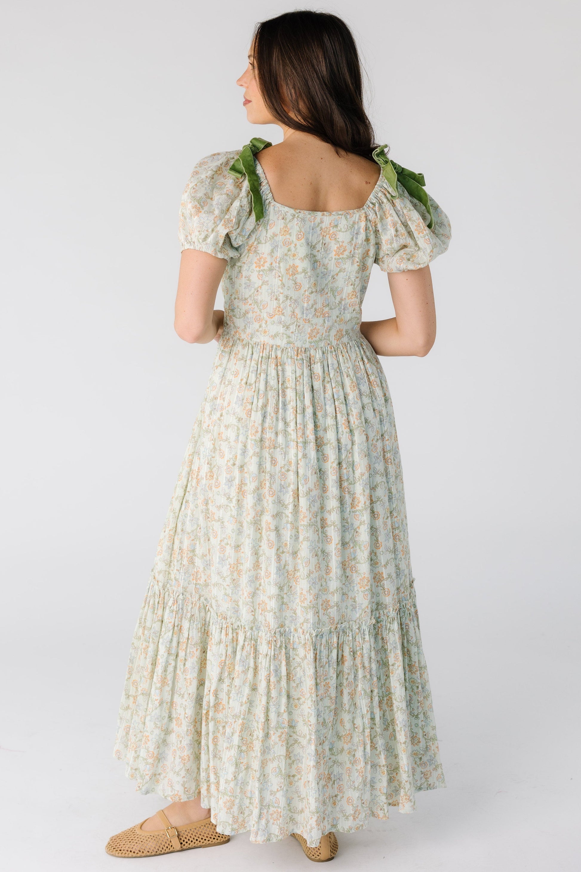 Back view of a woman wearing a green floral midi-maxi dress with a close fitting bodice and waist with full gathered skirt and short puff sleeves.