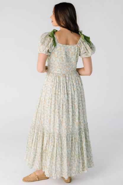 Back view of a woman wearing a green floral midi-maxi dress with a close fitting bodice and waist with full gathered skirt and short puff sleeves.