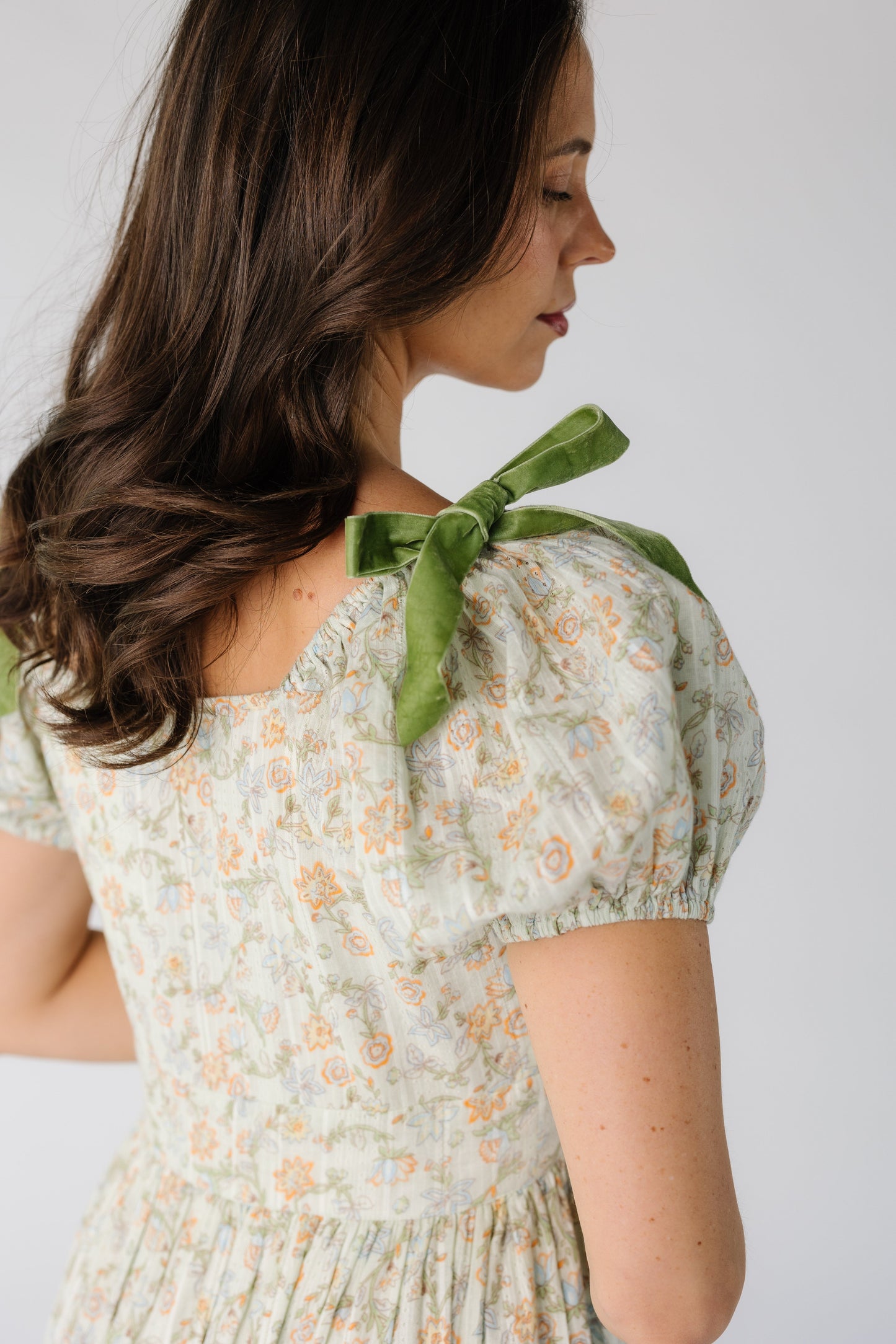 Back bodice view of a woman wearing a green floral print dress with a square back neckline, short puff sleeves and velvet bows on the shoulder.