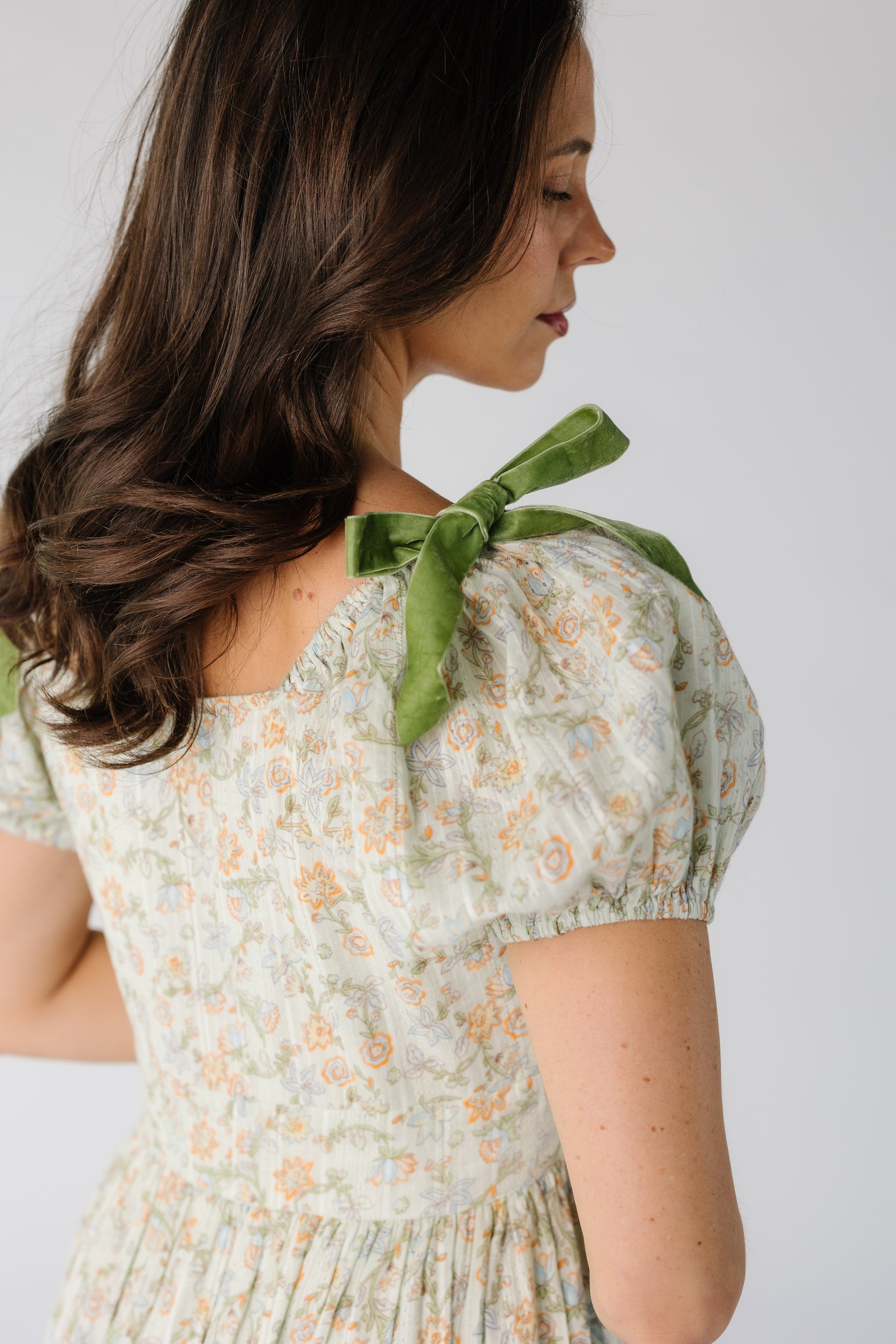Back bodice view of a woman wearing a green floral print dress with a square back neckline, short puff sleeves and velvet bows on the shoulder.