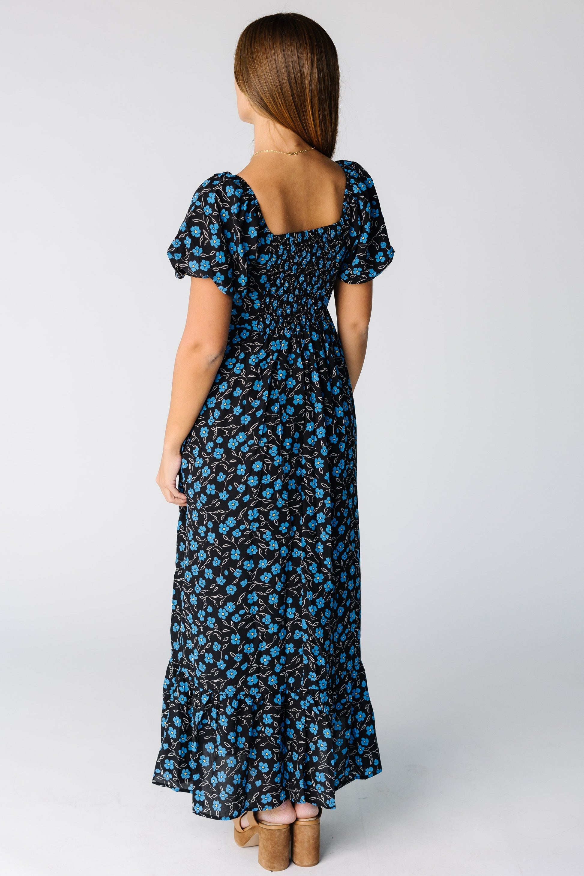 Back view of a woman wearing a blue floral maxi dress with a square back neckline and smocked back bodice for a snug fit