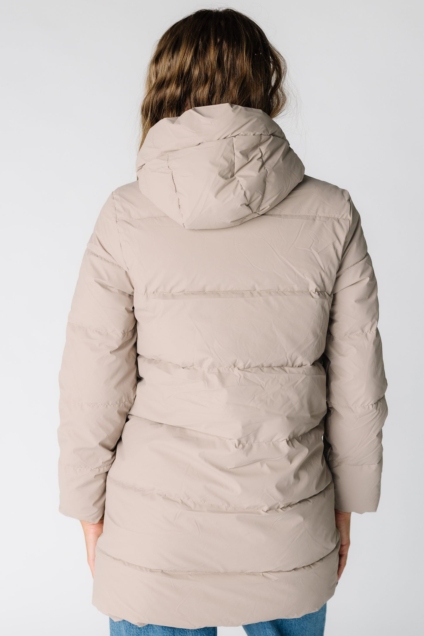 Back view of a woman wearing a taupe puffer jacket with long sleeves and a puffy hood