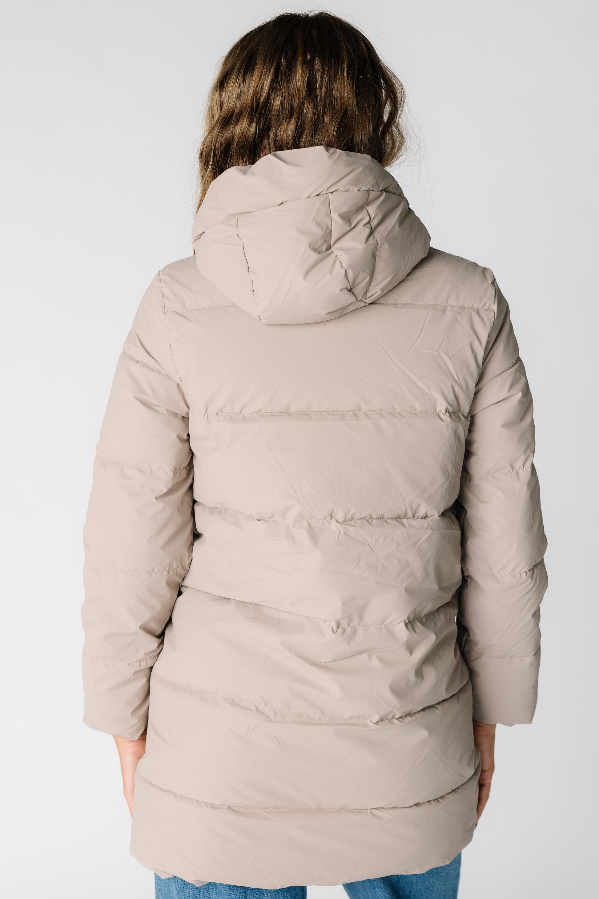Back view of a woman wearing a taupe puffer jacket with long sleeves and a puffy hood