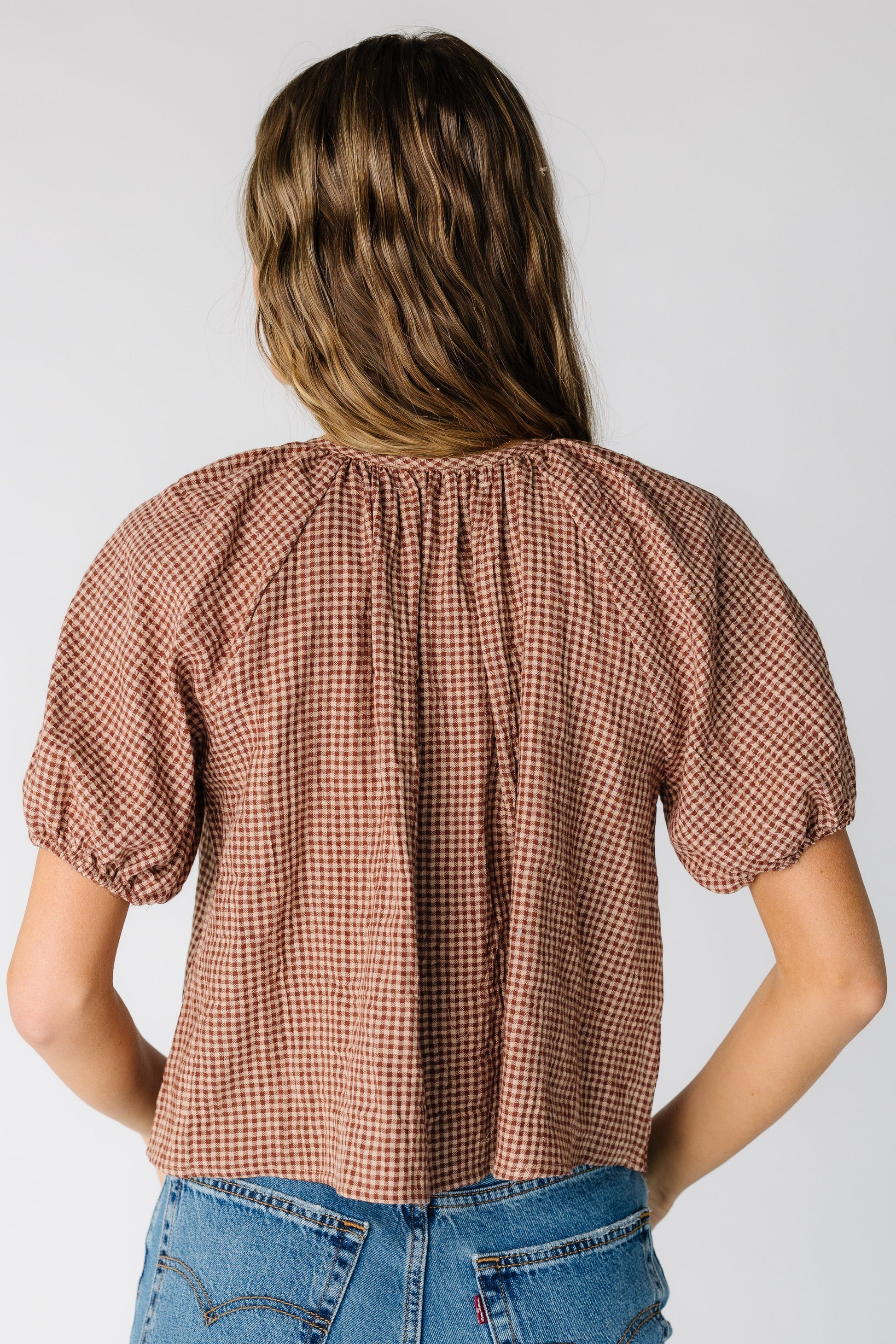 Back view of a woman wearing a loose fitting  taupe gingham top with short puff sleeves