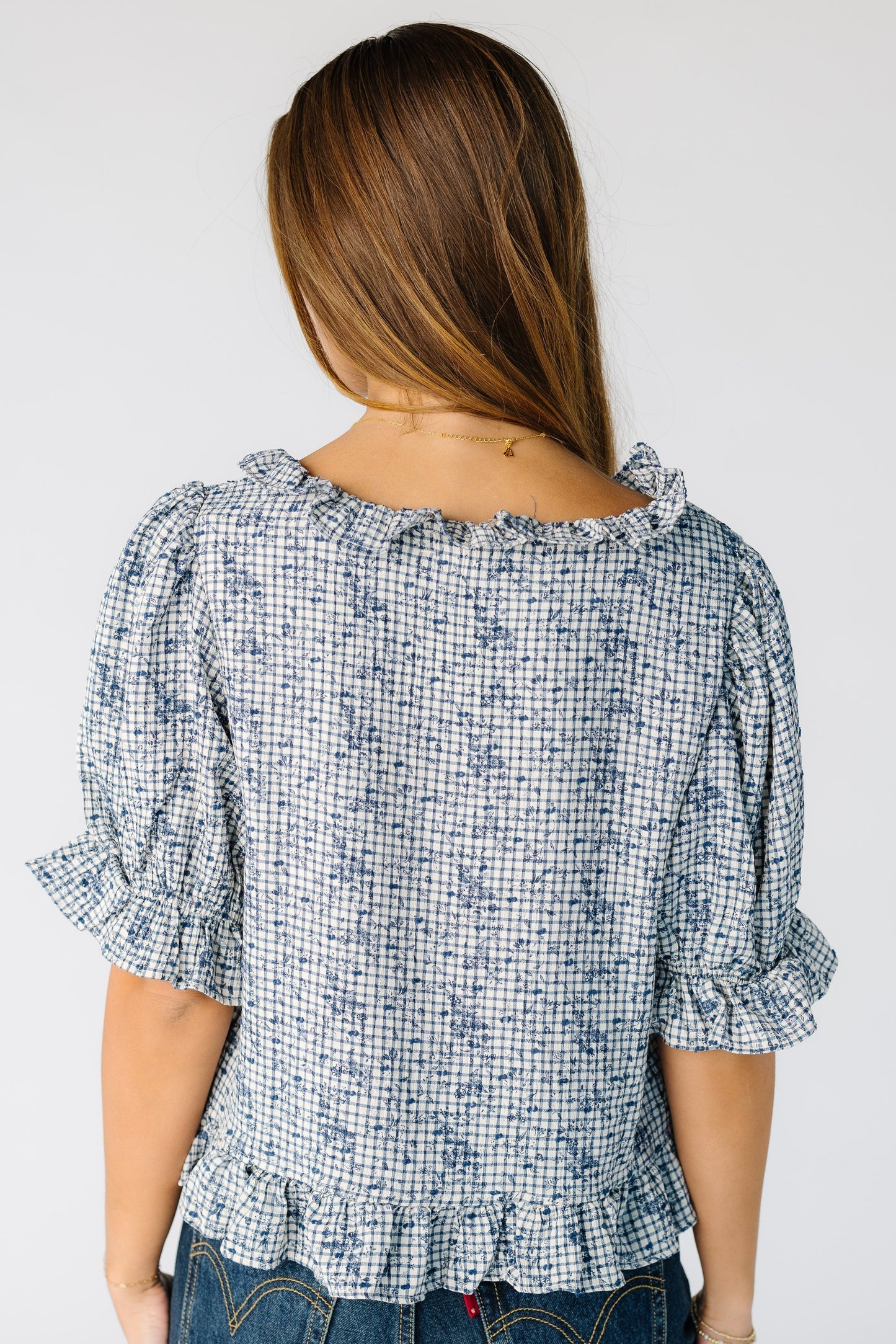 Back view of a woman wearing a loose  blue plaid top with a ruffle neckline with loose sleeves