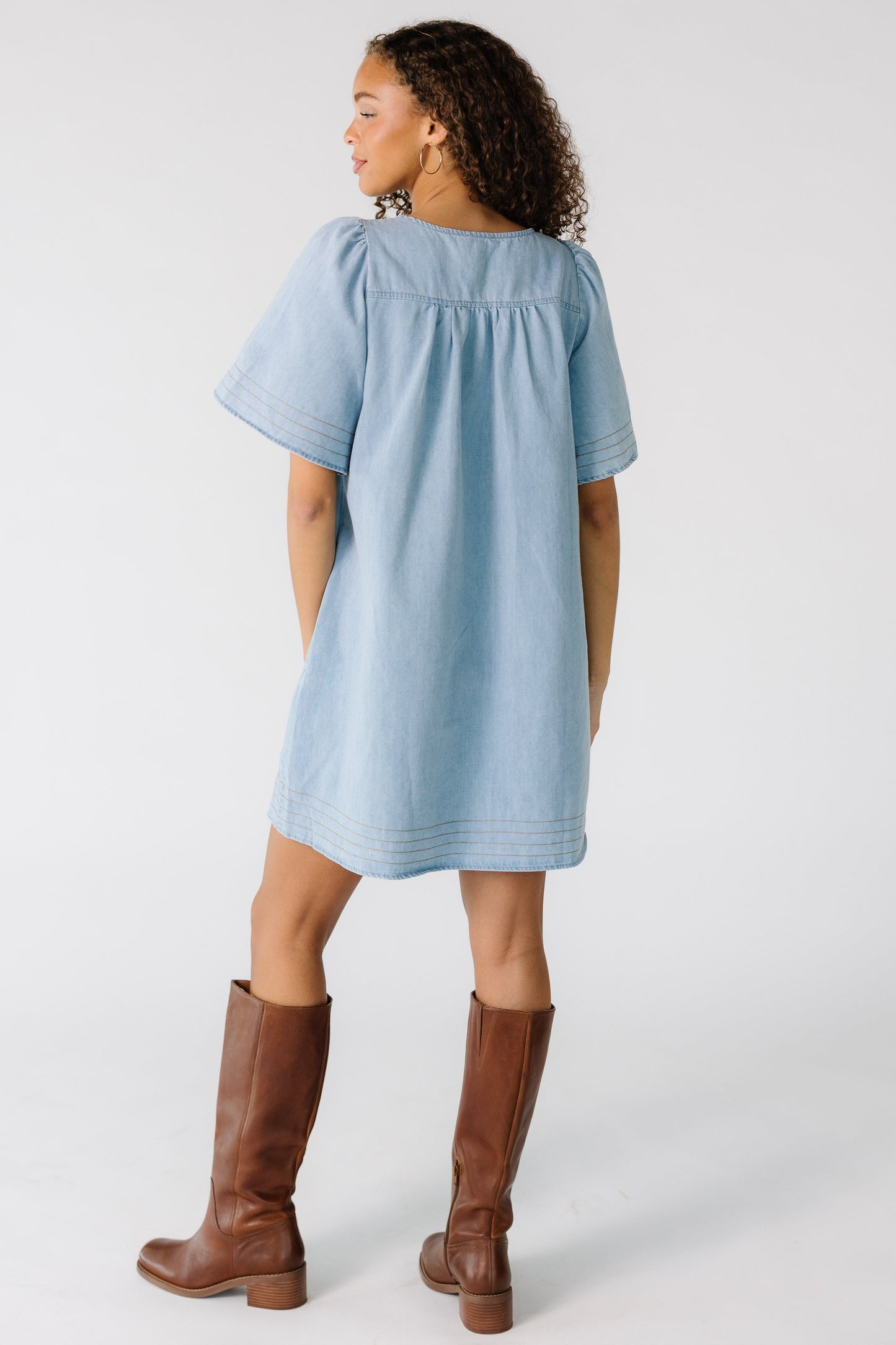 Back view of a teenage girl wearing a light denim short dress with short sleeves.