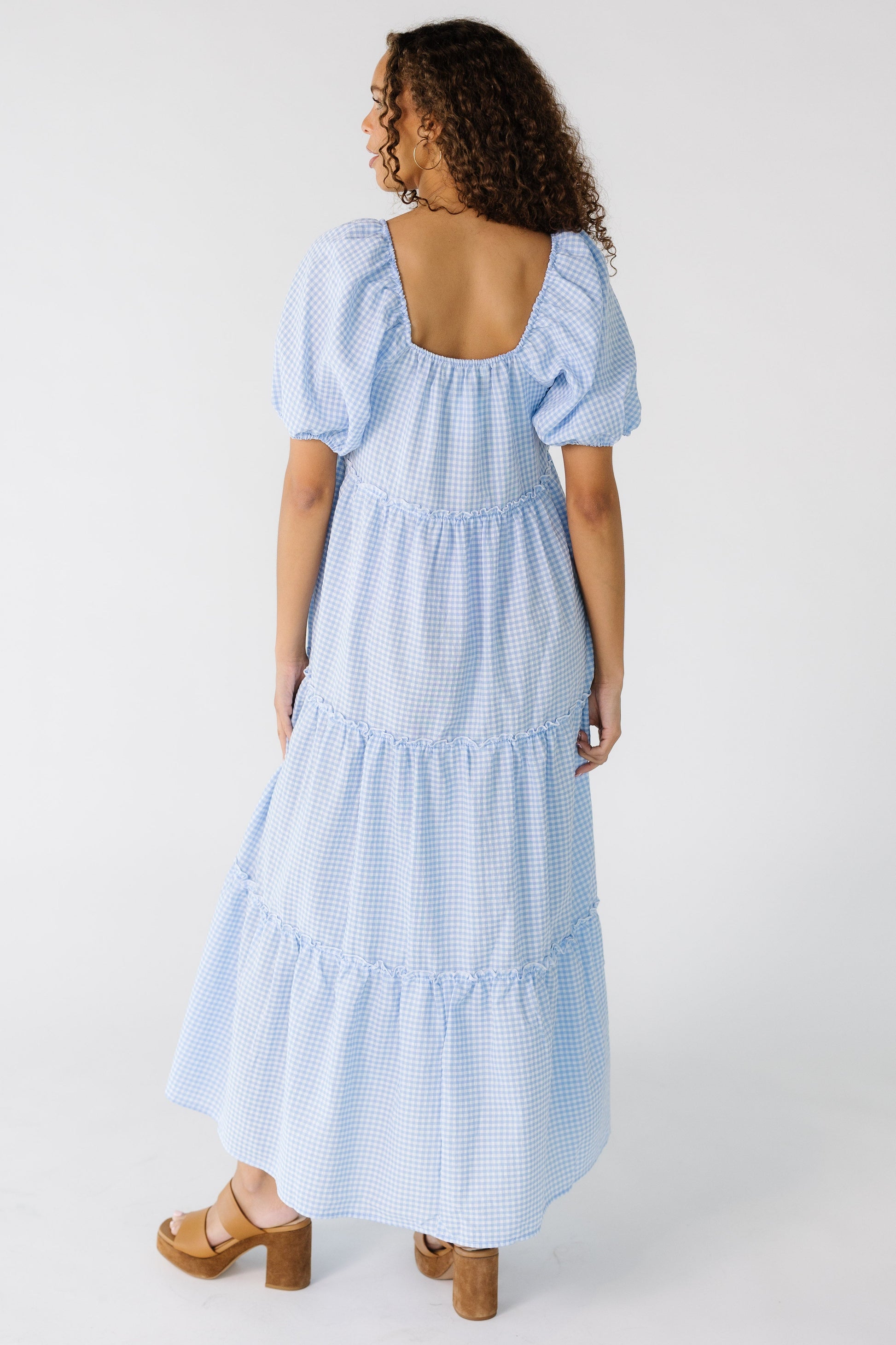 Back view of a  tee girl wearing a light blue gingham midi dress with a square back neckline and half length sleeves.