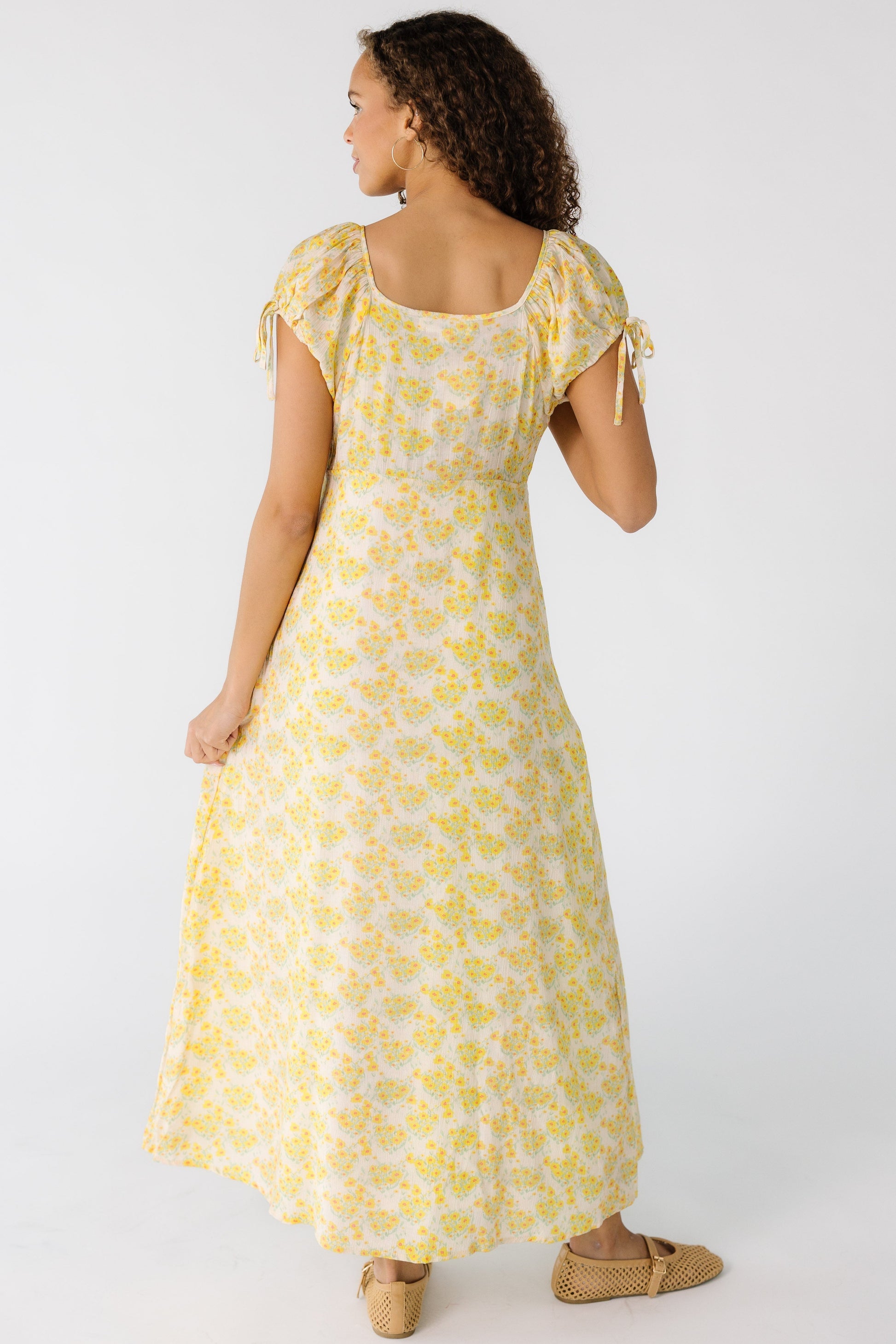 Back view of a teen girl wearing a yellow floral midi-maxi dress with a square back neckline and short puff sleeves.