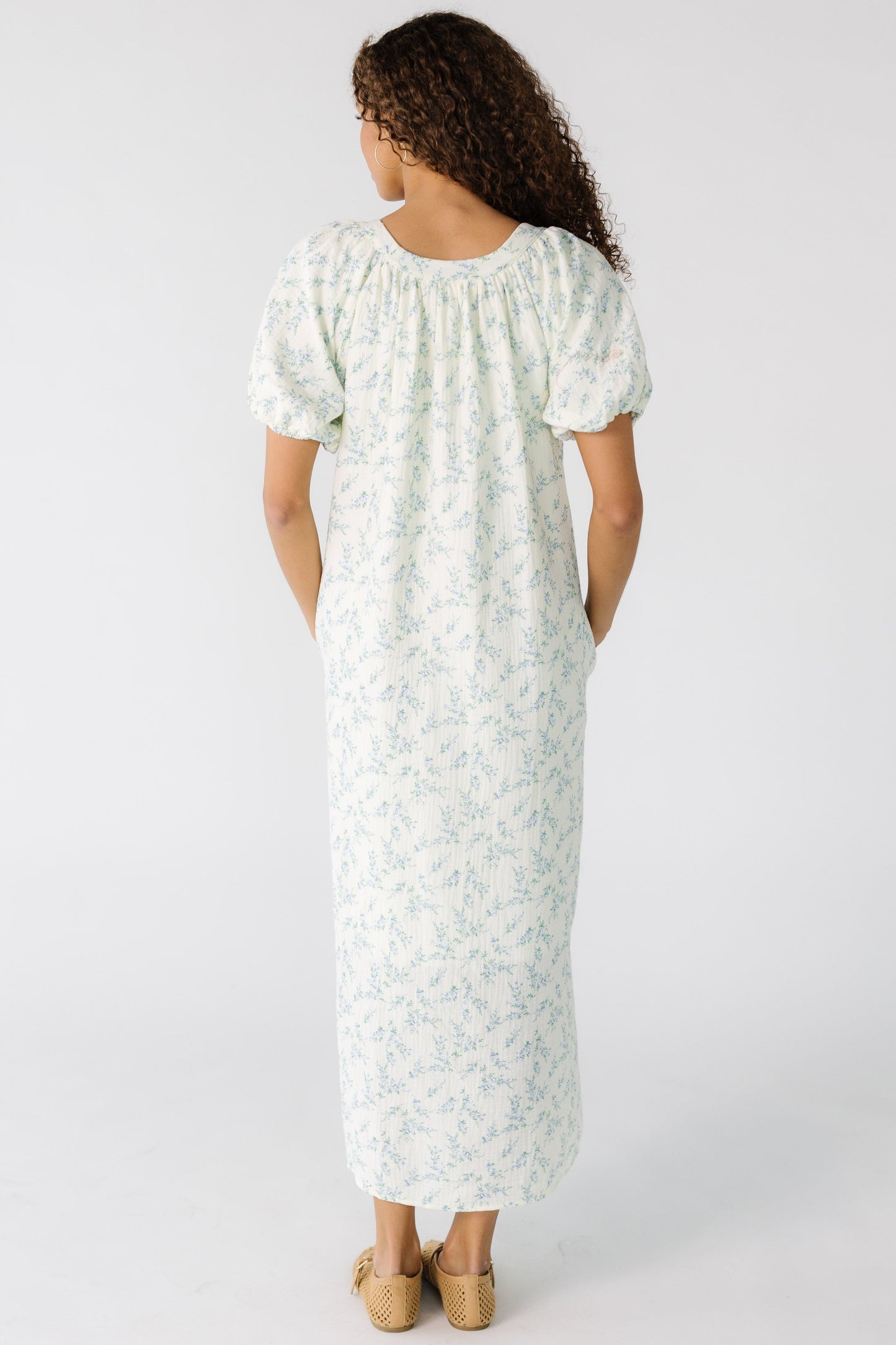 Back view of a teen girl wearing a cream midi dress with blue floral print with short puff sleeves.