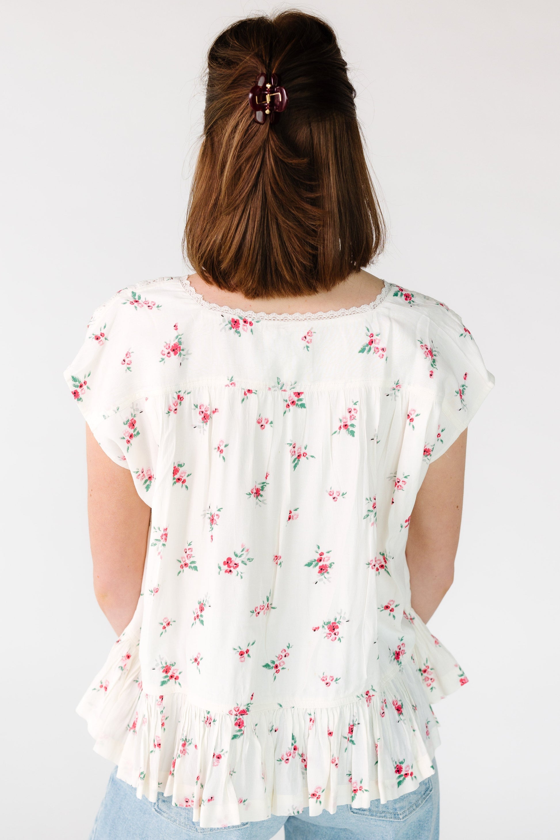 Back view of a woman wearing an off white floral, loose fitting blouse with a bottom ruffle.