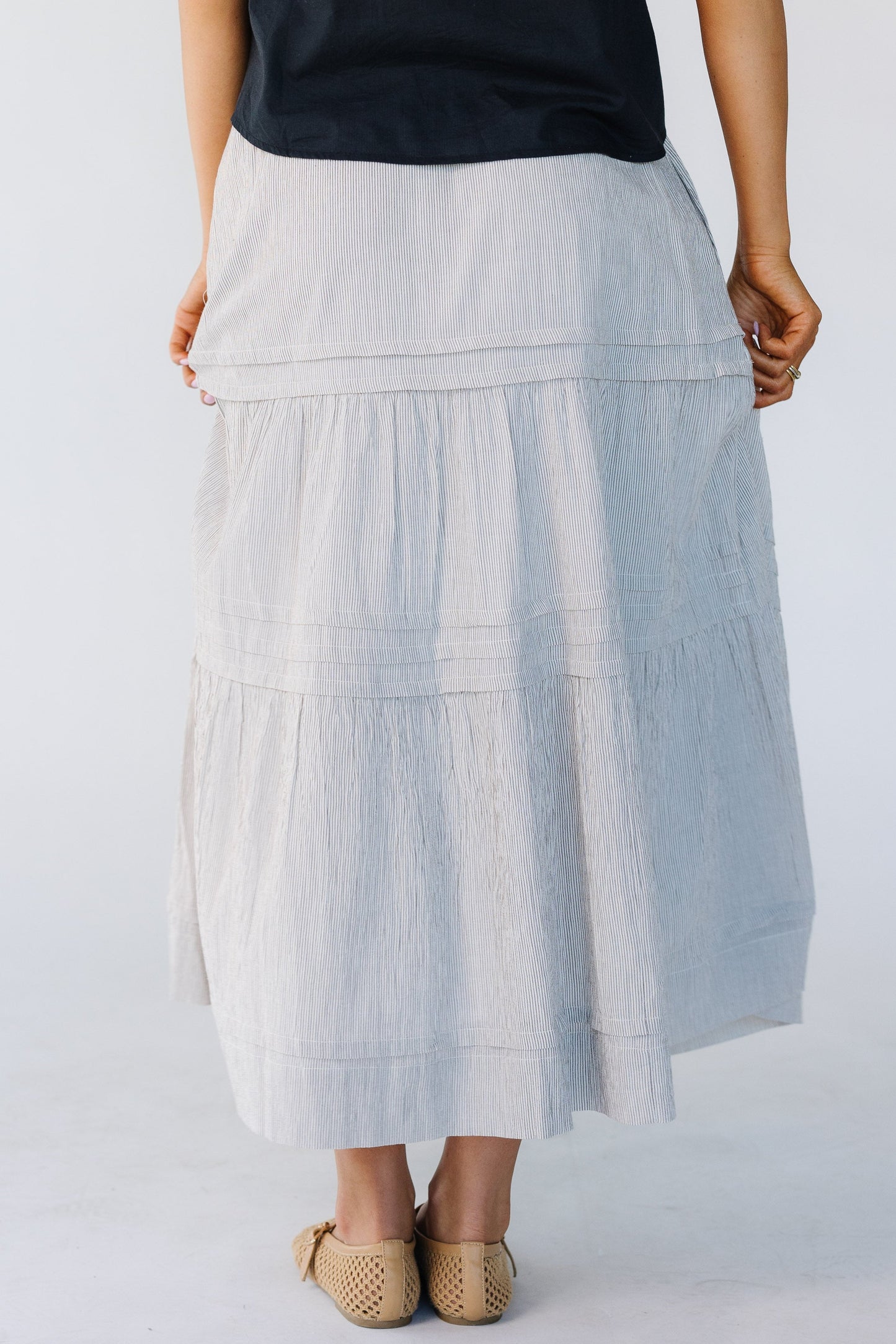 Back view of a woman wearing a narrow brown vertical stripe skirt with horizontal pleats and 2 gathered tiers.