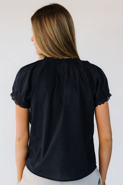 Back view of a woman wearing a black woven blouse with a lace trimmed round neckline ad short puff sleeves with a lace hem.