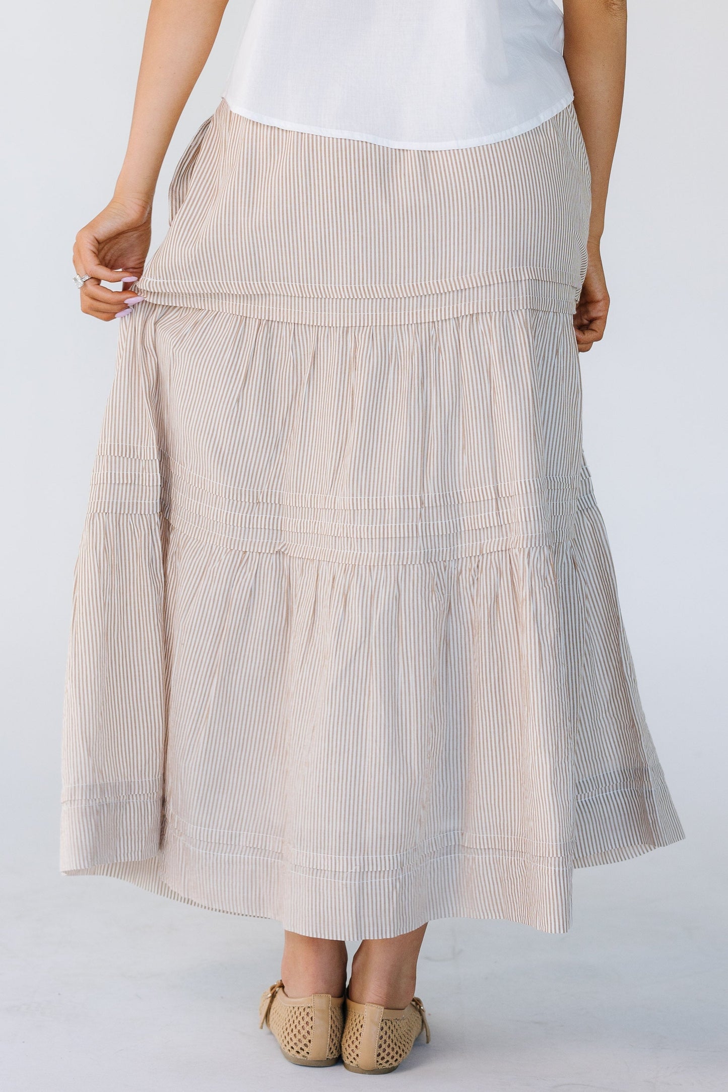 Back view of a woman wearing a  camel and white vertical stripe midi skirt with horizontal pleats and gathered tiers.