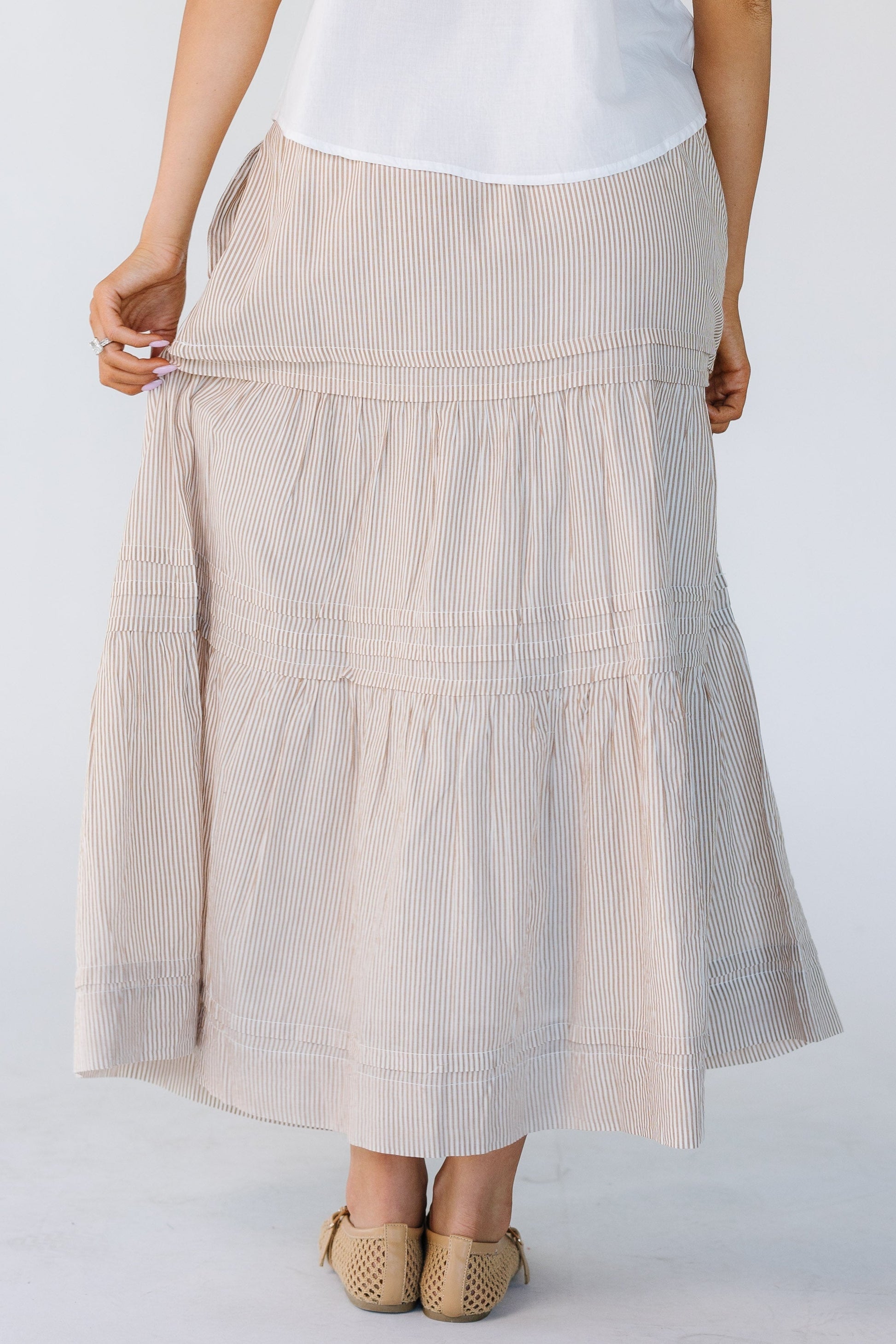 Back view of a woman wearing a  camel and white vertical stripe midi skirt with horizontal pleats and gathered tiers.