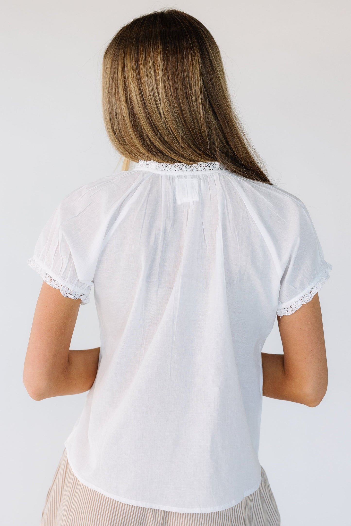Back view of a woman wearing a white blouse with short, lace hemmed sleeves and a high round neckline with lace trim.