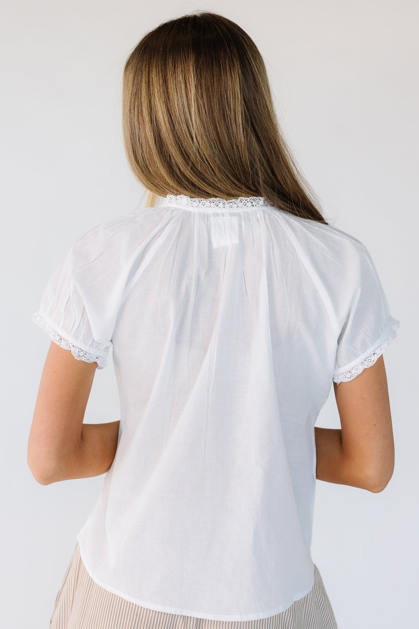 Back view of a woman wearing a white loose fitting blouse with lace trimmed short sleeves, and a lace trimmed neckline.