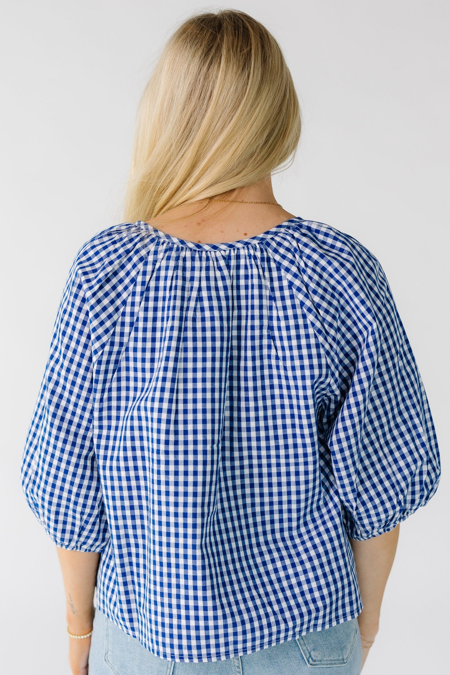 Back view of a woman wearing a loose fitting blue and white gingham top with a round neckline and half length sleeves.