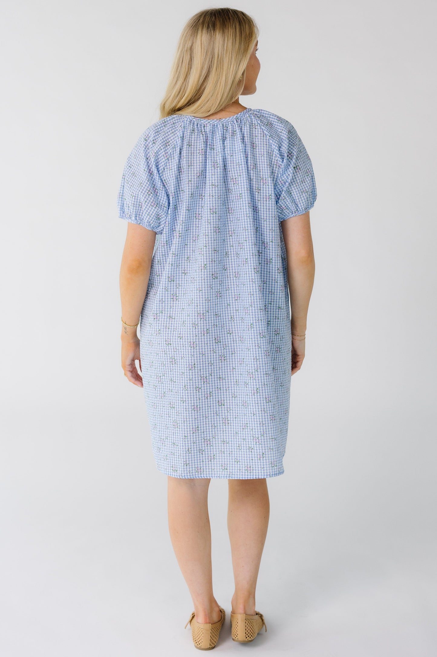 Back view of a woman wearing a  light blue gingham knee length dress with short sleeves and a round neckline.