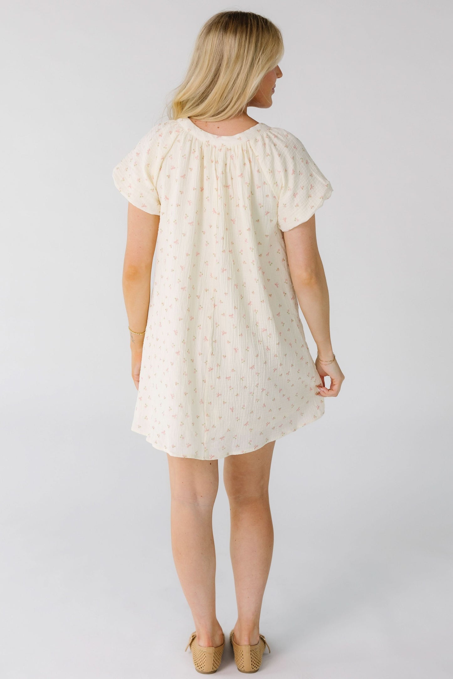 Back view of a woman wearing a cream with pink print mini dress with short balloon sleeves.