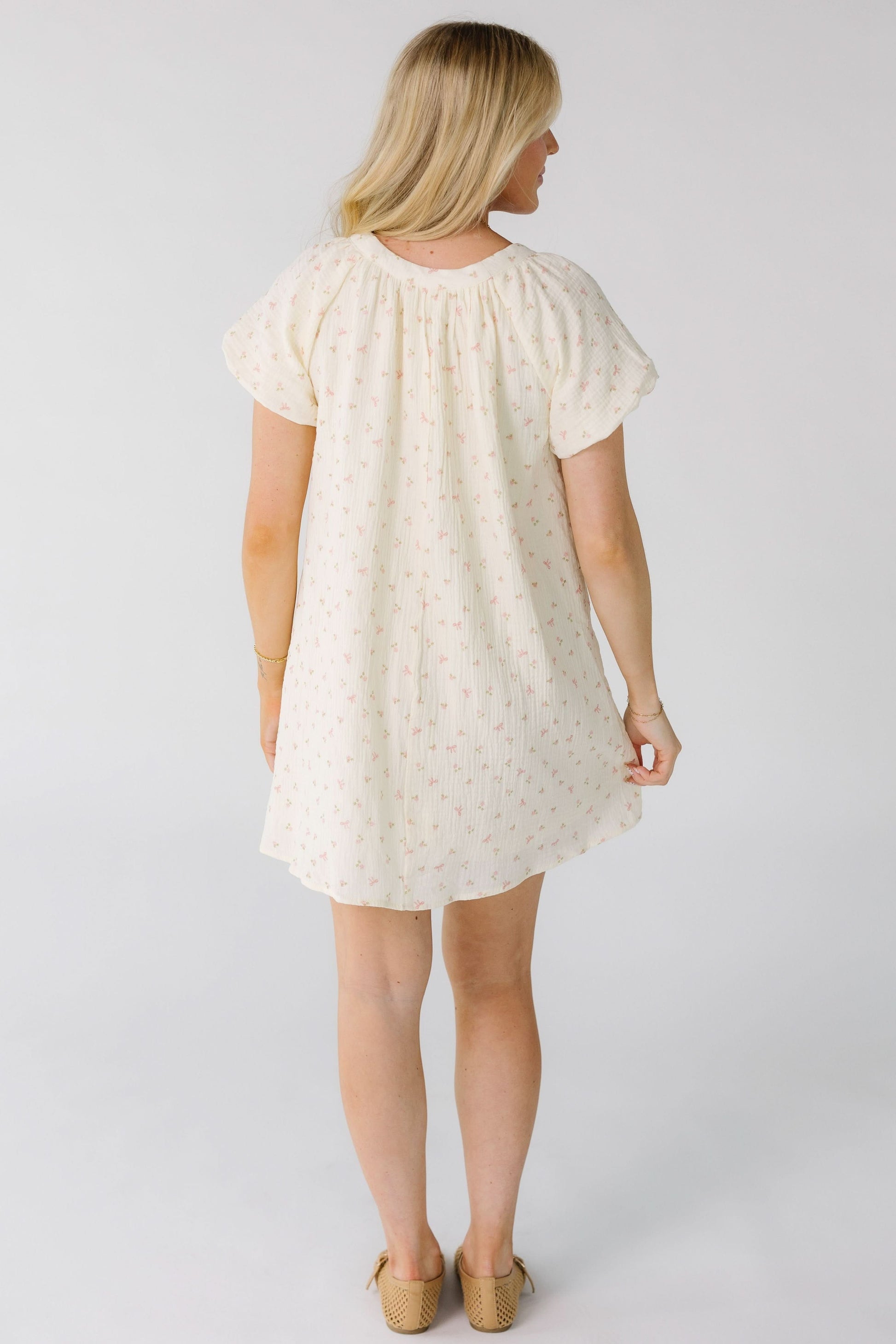 Back view of a woman wearing a cream with pink print mini dress with short balloon sleeves.