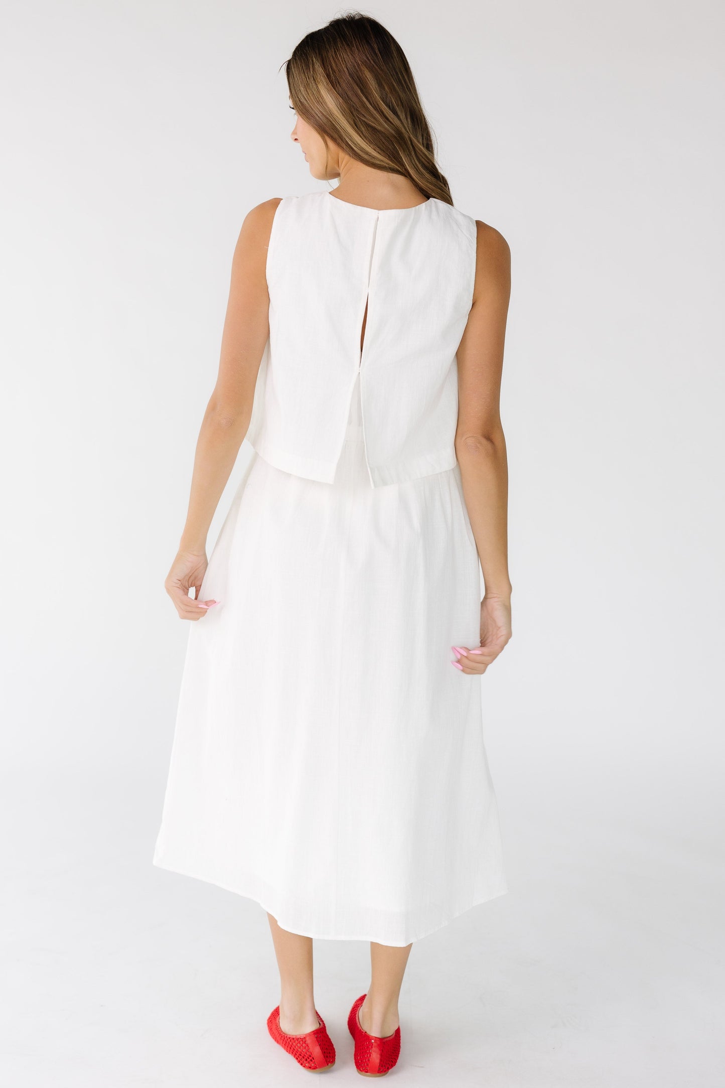 Back view of a woman wearing a sleeveless 1 piece dress hat looks like a loose fitting top with a gathered midi skirt in off white.