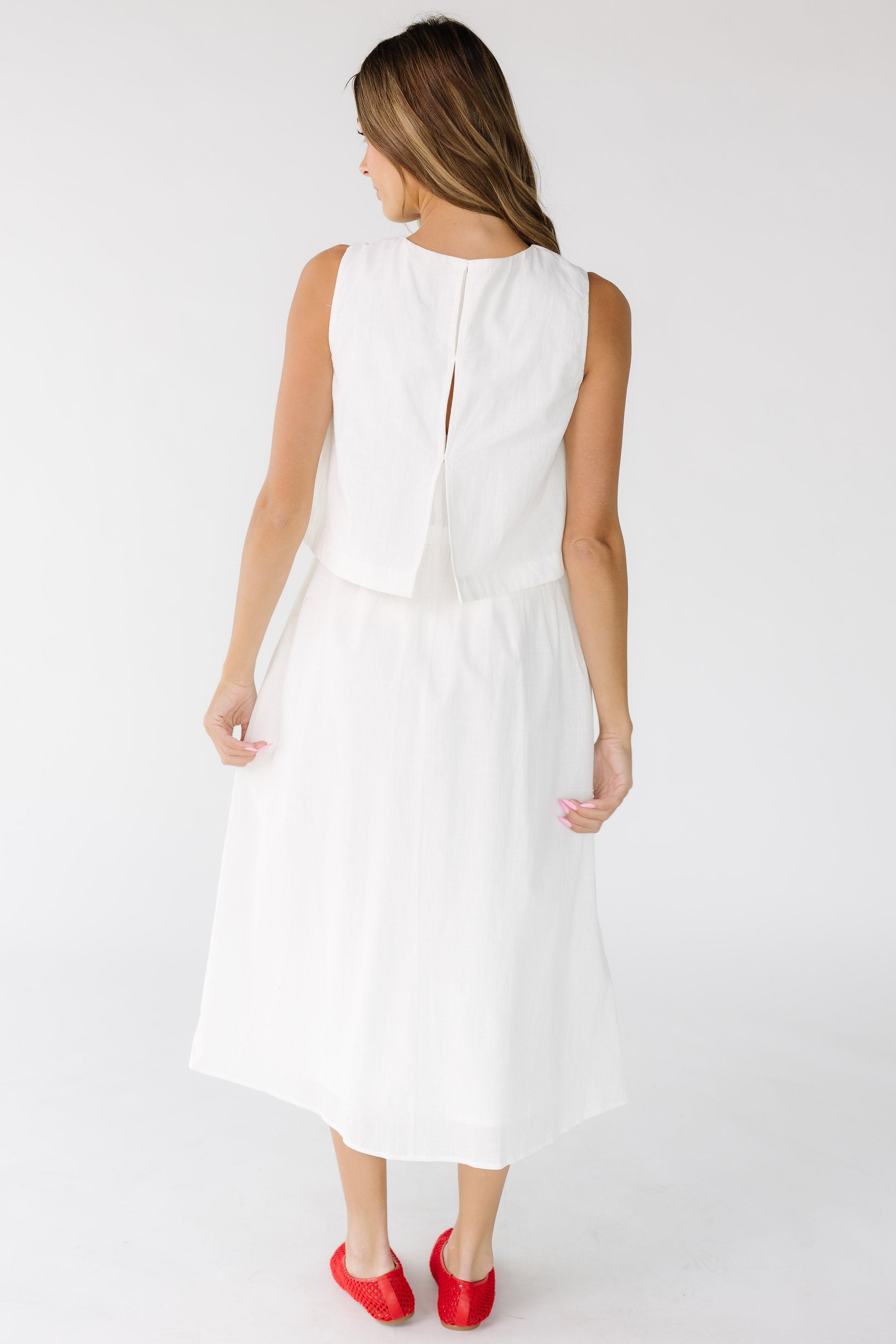 Back view of a woman wearing a sleeveless 1 piece dress hat looks like a loose fitting top with a gathered midi skirt in off white.