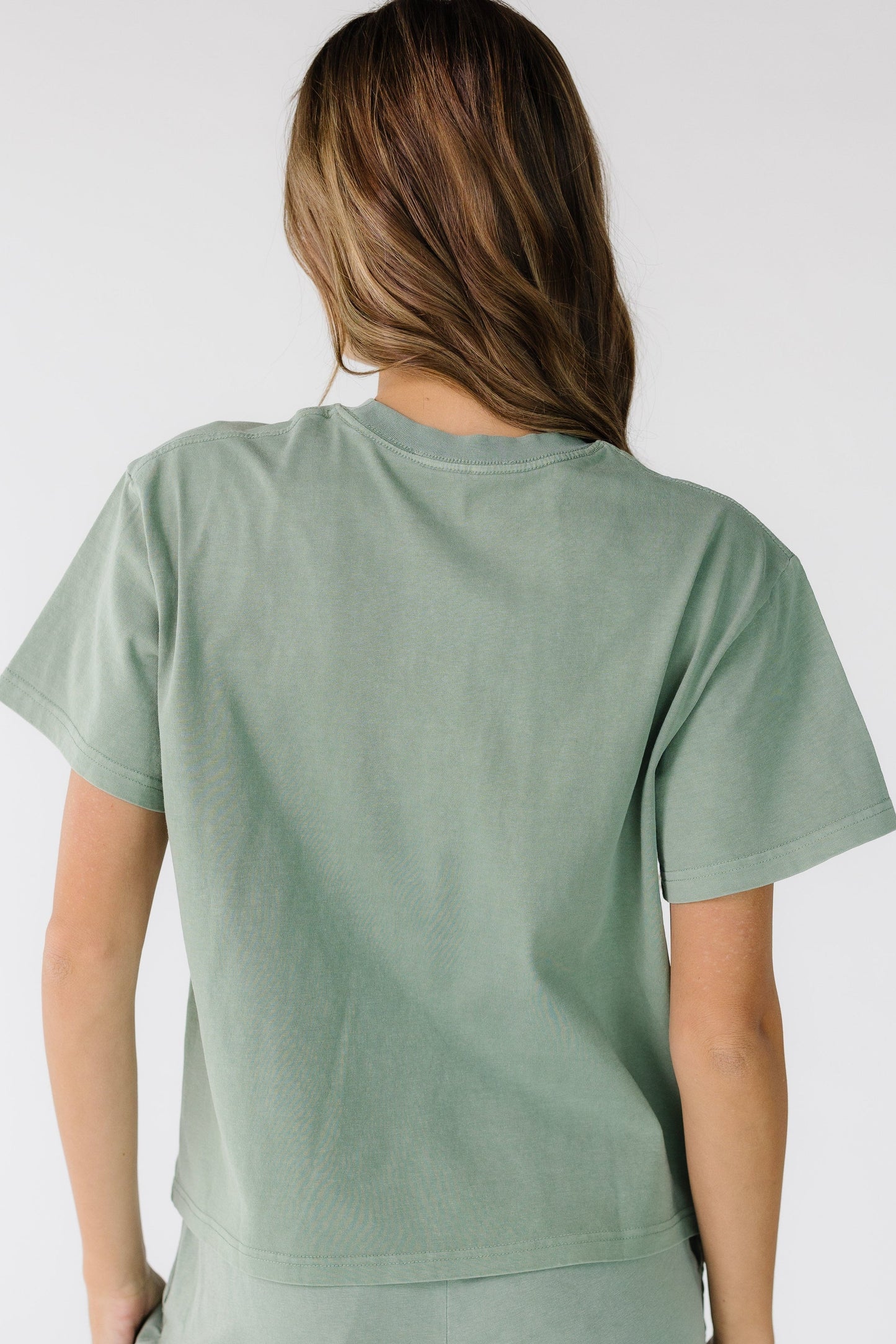 Back view of a woman wearing a sage green top with short sleeves and a crew neckline.