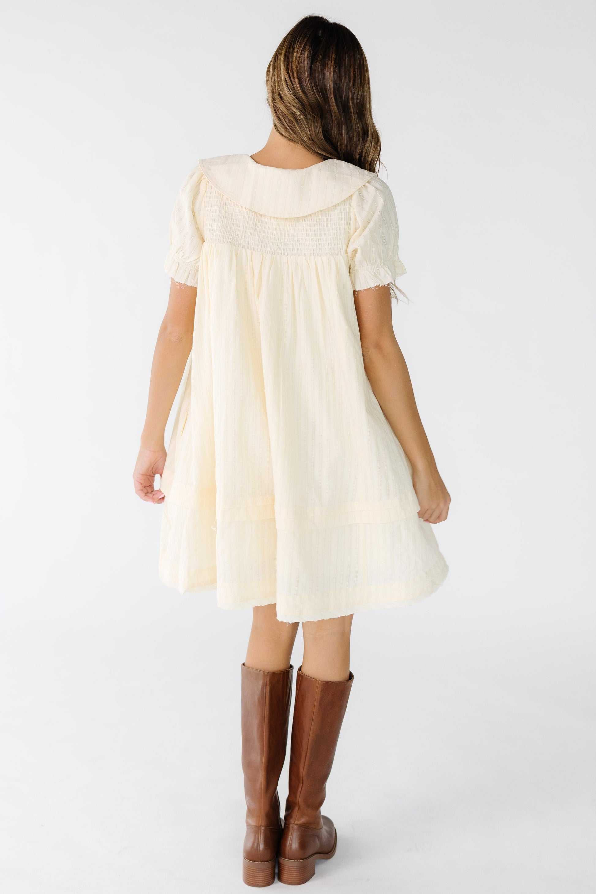 Back view of a woman wearing a cream short dress with smocked front and back yoke with short puff sleeves.